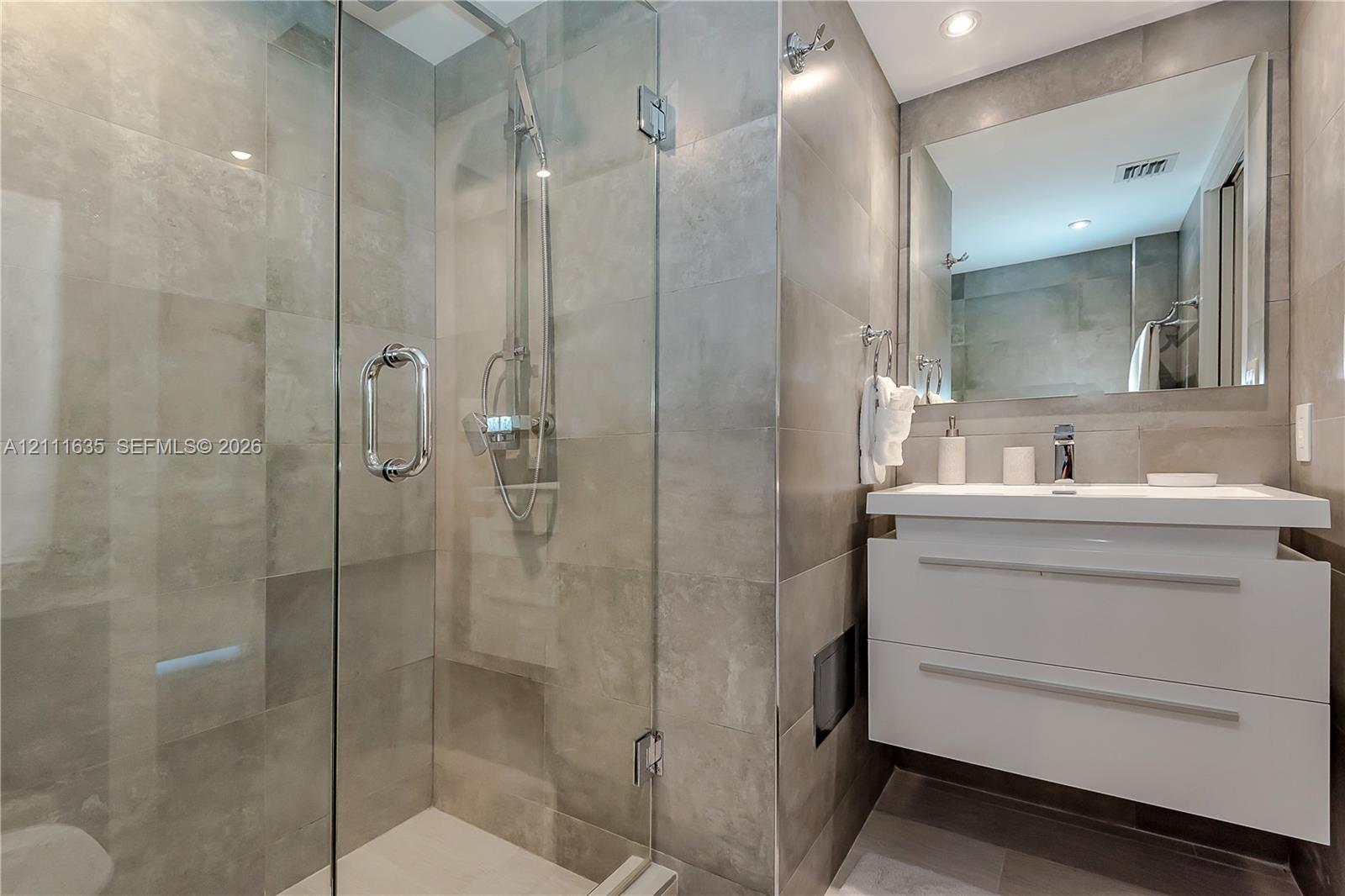 Walk-in shower with a glass enclosure, wall-mounted floating vanity, large rectangular mirror, and full-height stone-finish tile surround