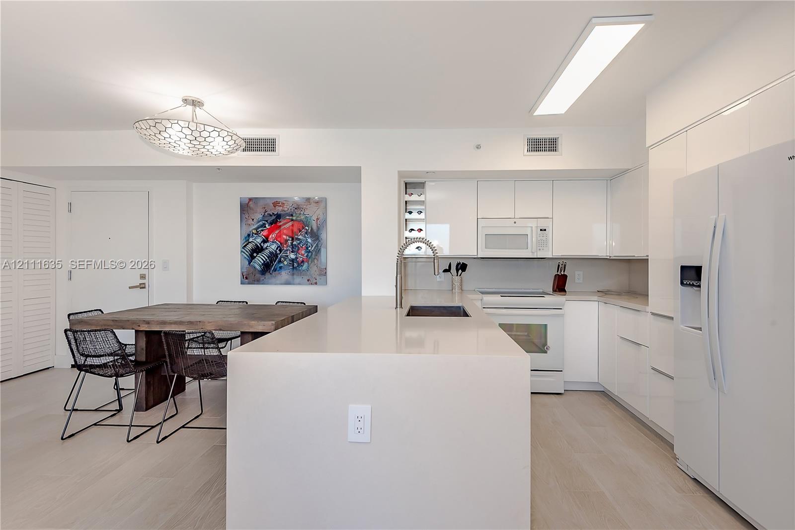 Kitchen featuring white slab cabinetry, an oversized quartz waterfall island, built-in wine storage, light-toned wood-finish flooring, and a modern geometric pendant light fixture
