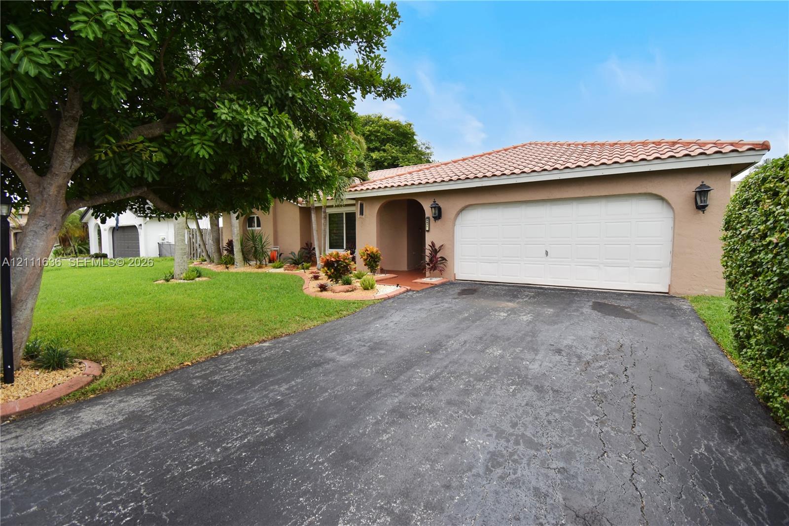 Stucco exterior featuring a clay tile roof, two-car garage, manicured lawn, and mature trees