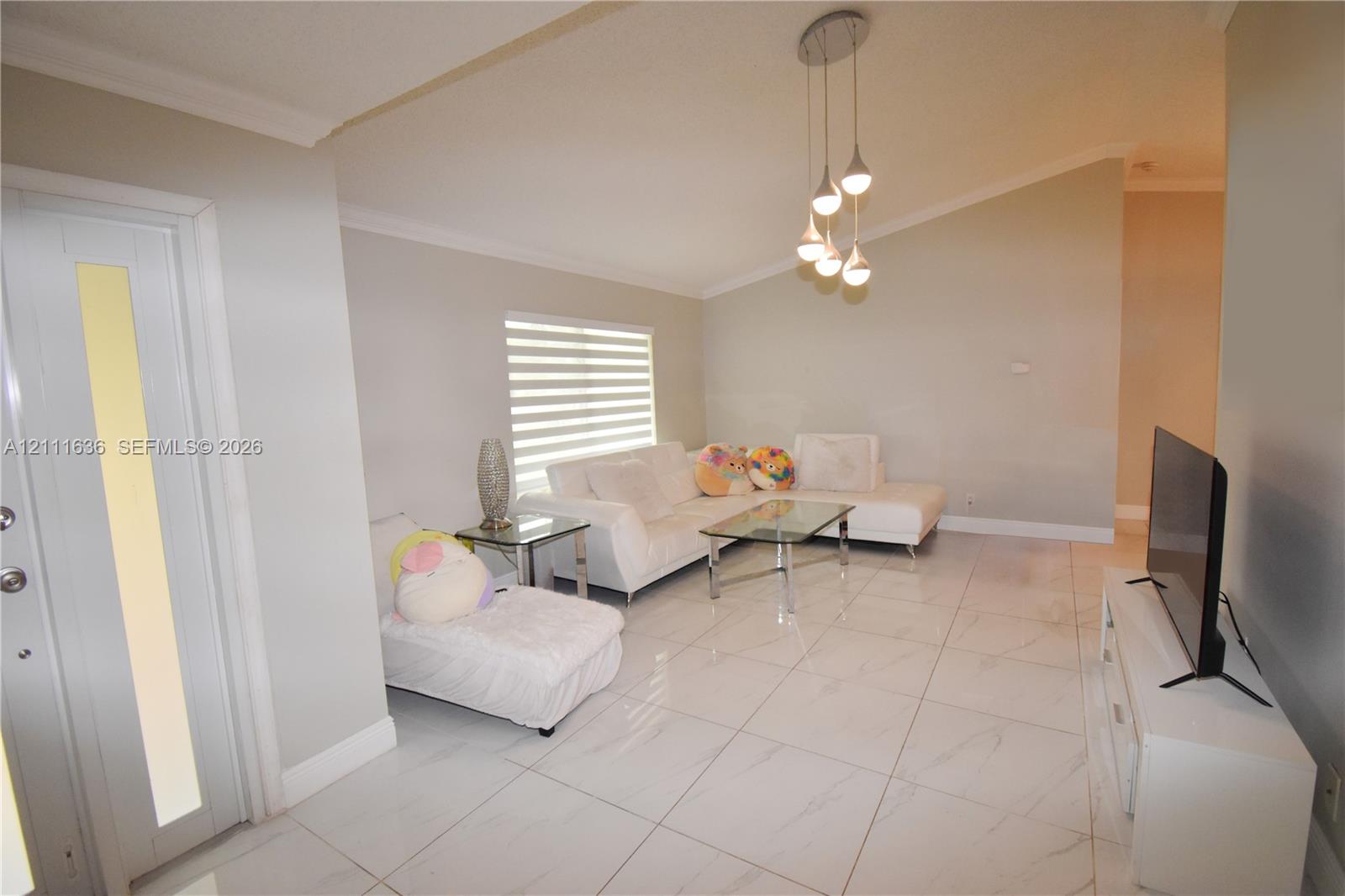 High-ceiling living area featuring large-format polished tile flooring, tiered multi-light pendant fixture, window with horizontal blinds, and glass-paneled entry door