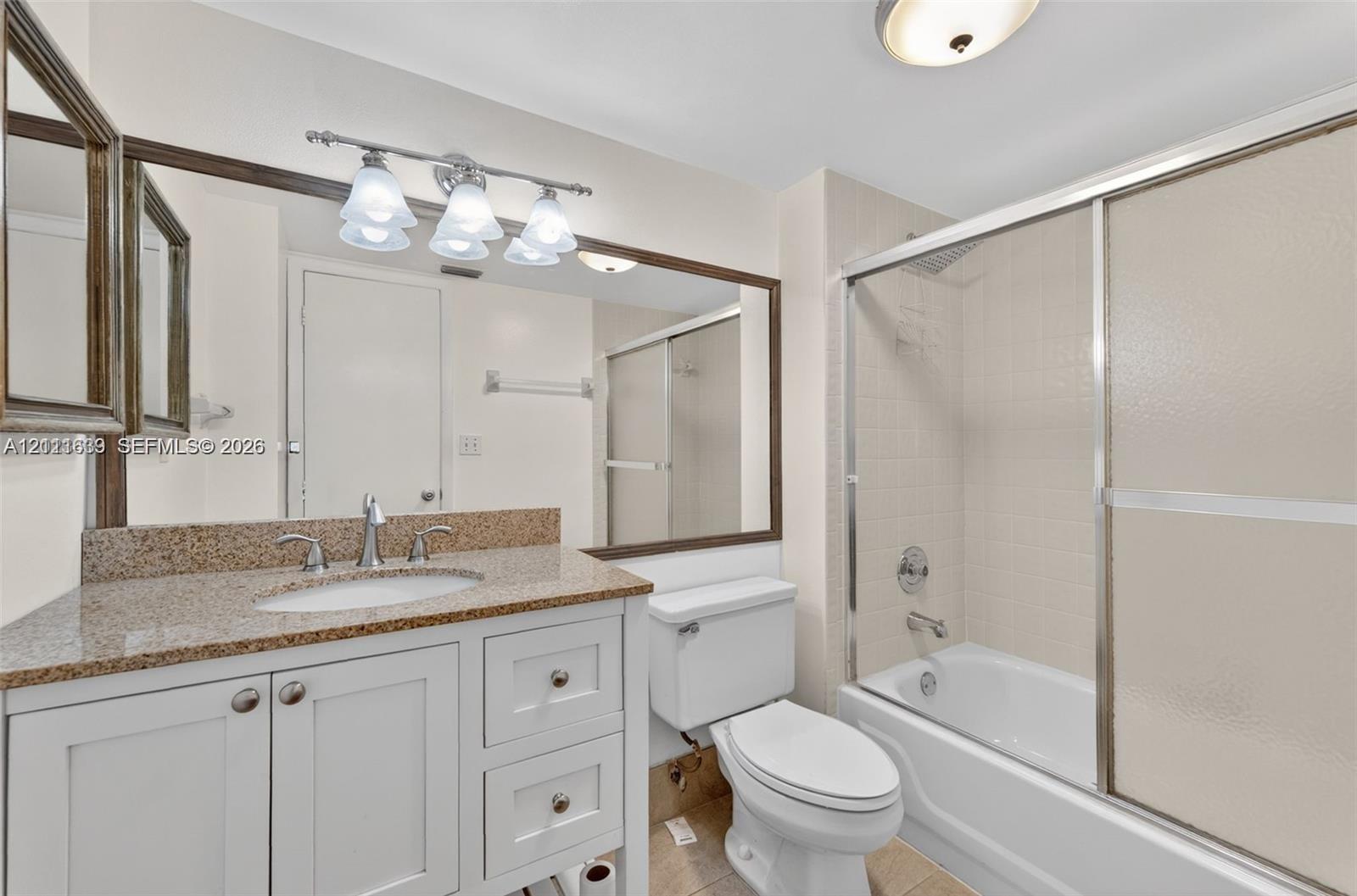 Bathroom featuring a white vanity with a stone countertop and integrated sink, a large wall-mounted mirror, a three-light fixture, and a shower-tub combination with sliding glass doors Bathroom featuring a white vanity with a stone countertop and integrated sink, a large wall-mounted mirror, a three-light fixture, and a shower-tub combination with sliding glass doors