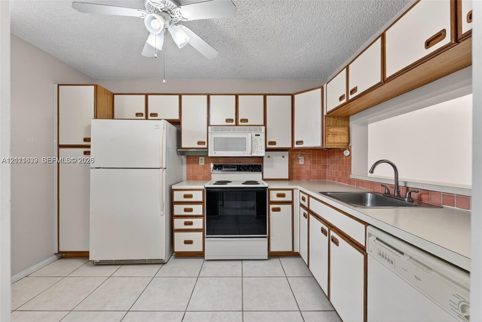 Kitchen featuring white cabinetry with wood trim, ceramic tile backsplash, integrated microwave, stainless steel sink, and tile flooring