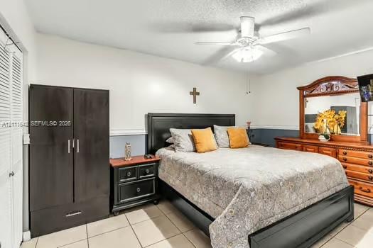 Bedroom featuring neutral-toned tile flooring, dual-door closet with louvered panels, and a central ceiling fan with integrated lighting