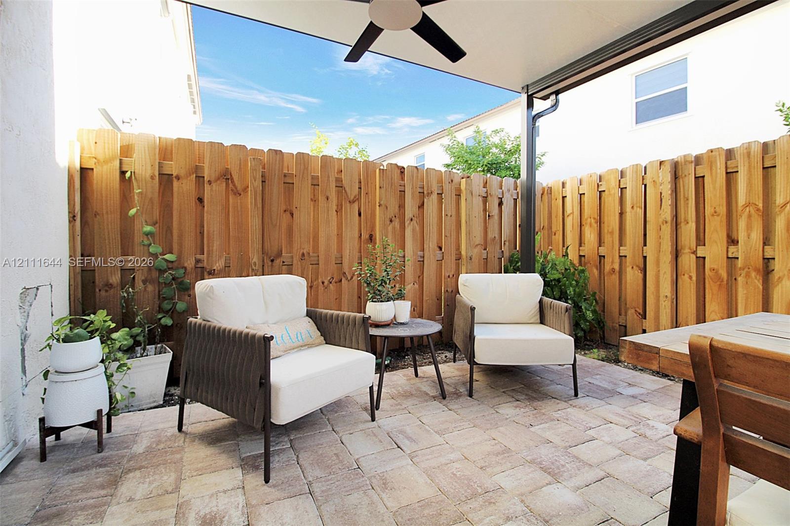 Covered outdoor patio featuring stone-paver flooring, a ceiling-mounted fan, and natural wood privacy fencing