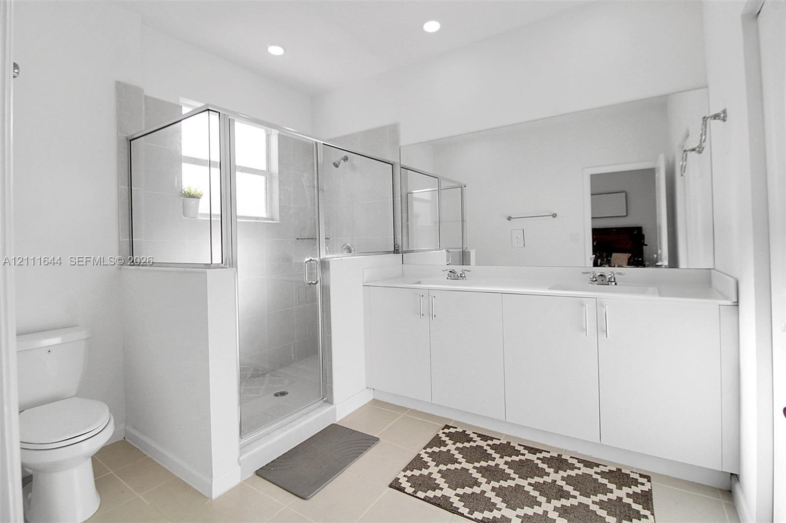 Double vanity with white cabinetry and solid surface countertop, large wall-mounted mirror, and glass-enclosed shower with tiled surround