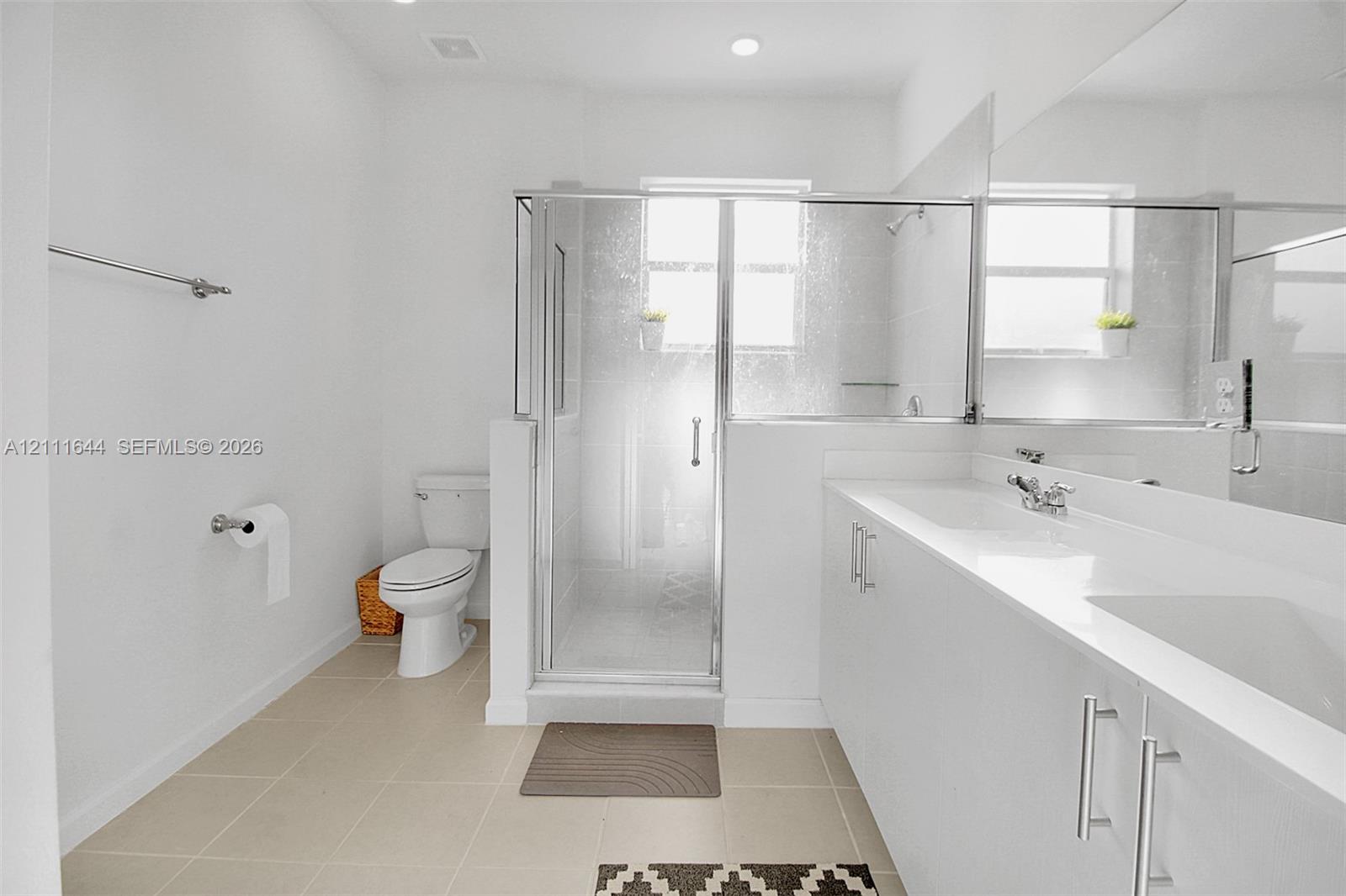 Dual-sink vanity with white cabinetry and solid surface countertop, glass-enclosed shower, polished chrome fixtures, and neutral floor tiling