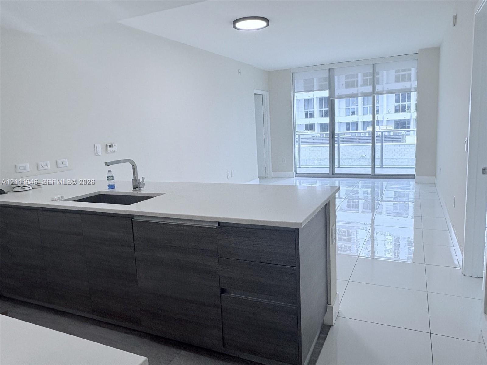 Kitchen island featuring dark wood-finish cabinetry, a white countertop, and an undermount sink with a gooseneck faucet, set against white tile flooring and floor-to-ceiling glass paneling