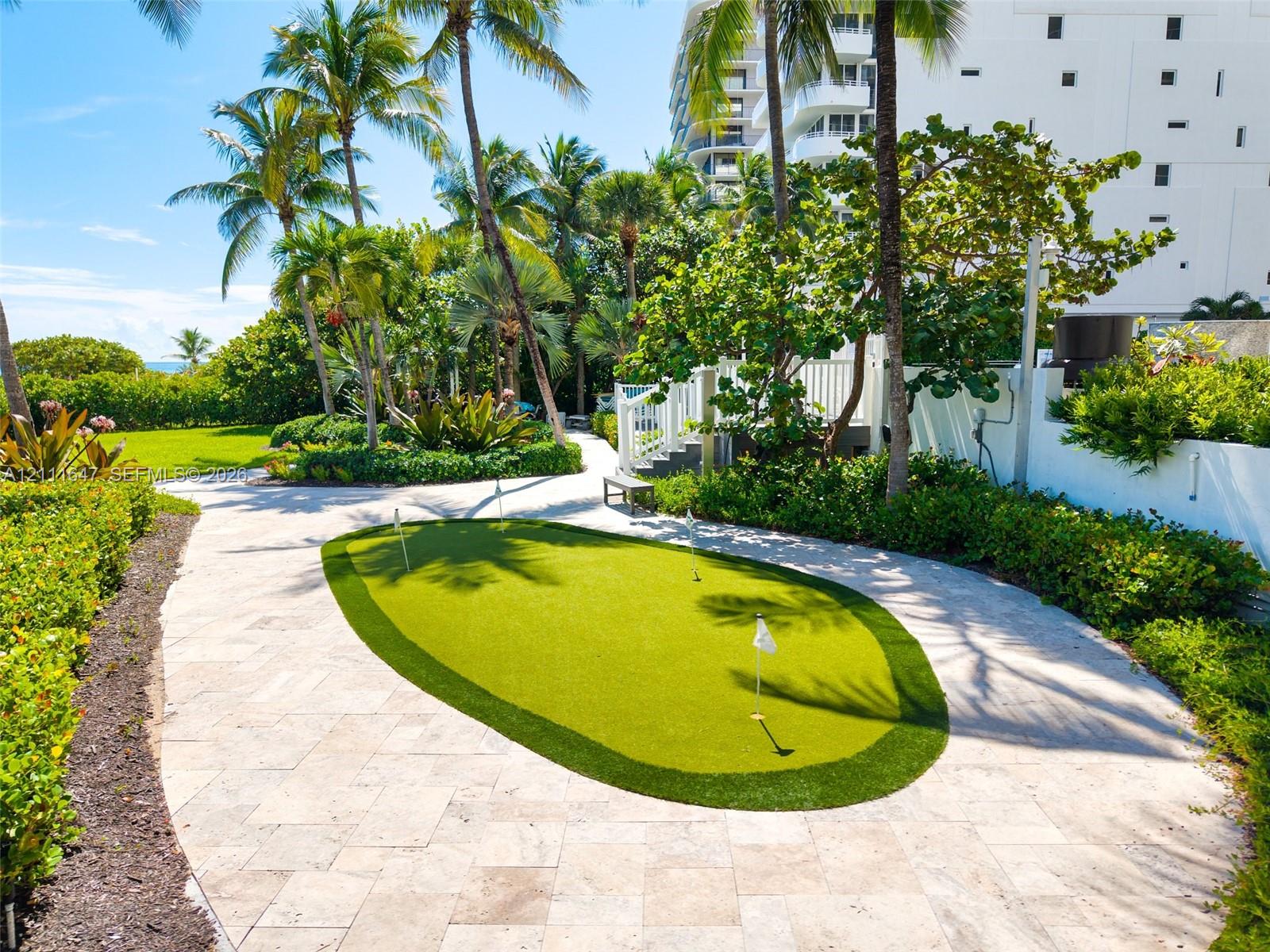 Putting green with synthetic turf surrounded by natural stone paver hardscaping, mature palm trees, and landscaped gardens