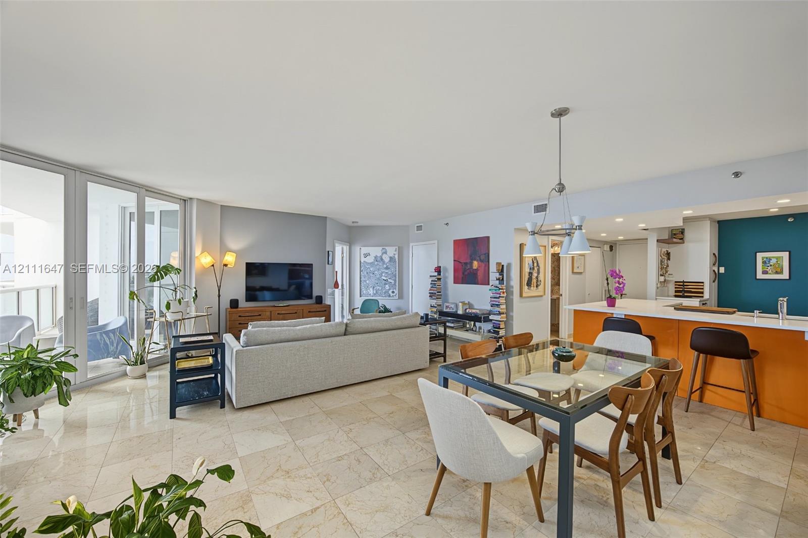 Open-concept interior featuring light-toned tile flooring, floor-to-ceiling glass sliding doors, recessed lighting, a built-in kitchen peninsula, and a multi-pendant light fixture