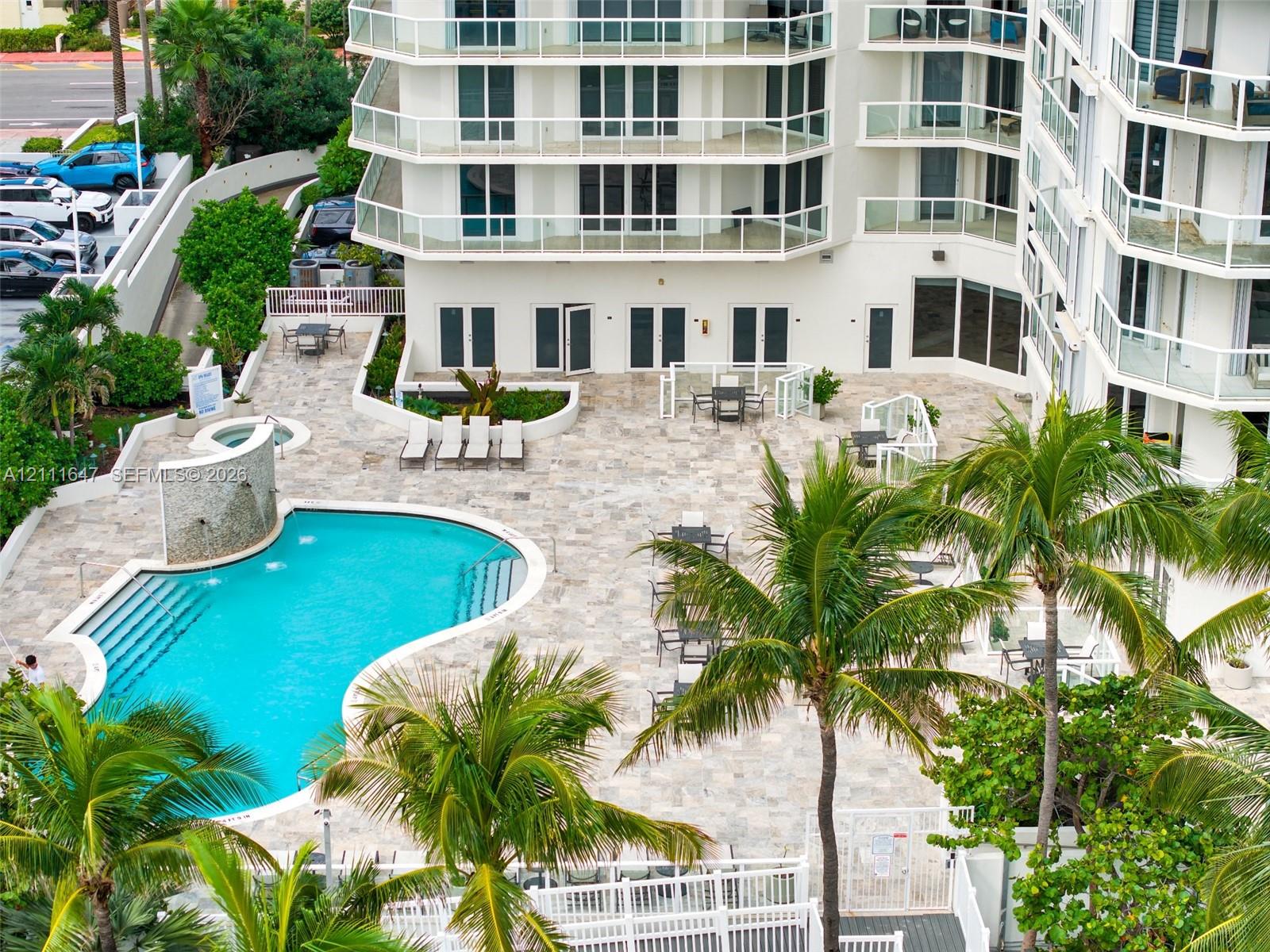 Multi-story building featuring tiered balconies with glass railings, expansive stone-paved pool deck, swimming pool with integrated spa and water feature, and established tropical landscaping