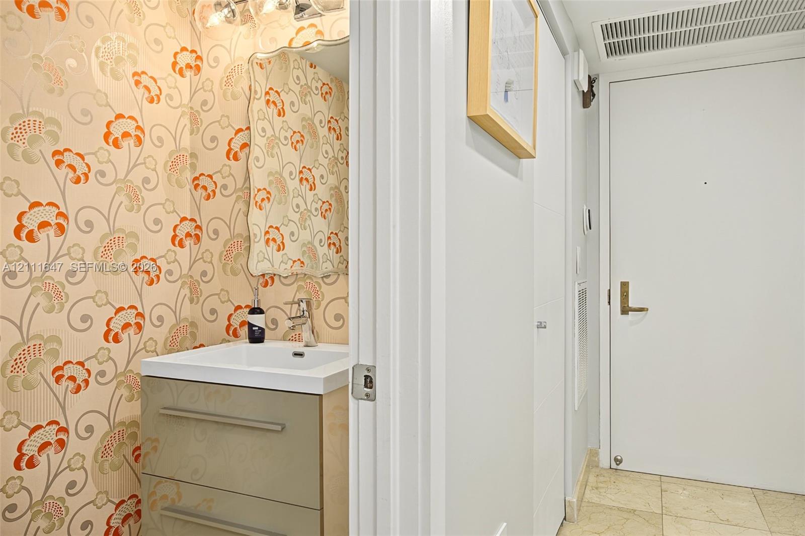 Powder room featuring floral patterned wallcovering, wall-mounted mirror, two-drawer vanity with a white basin, and polished metal hardware