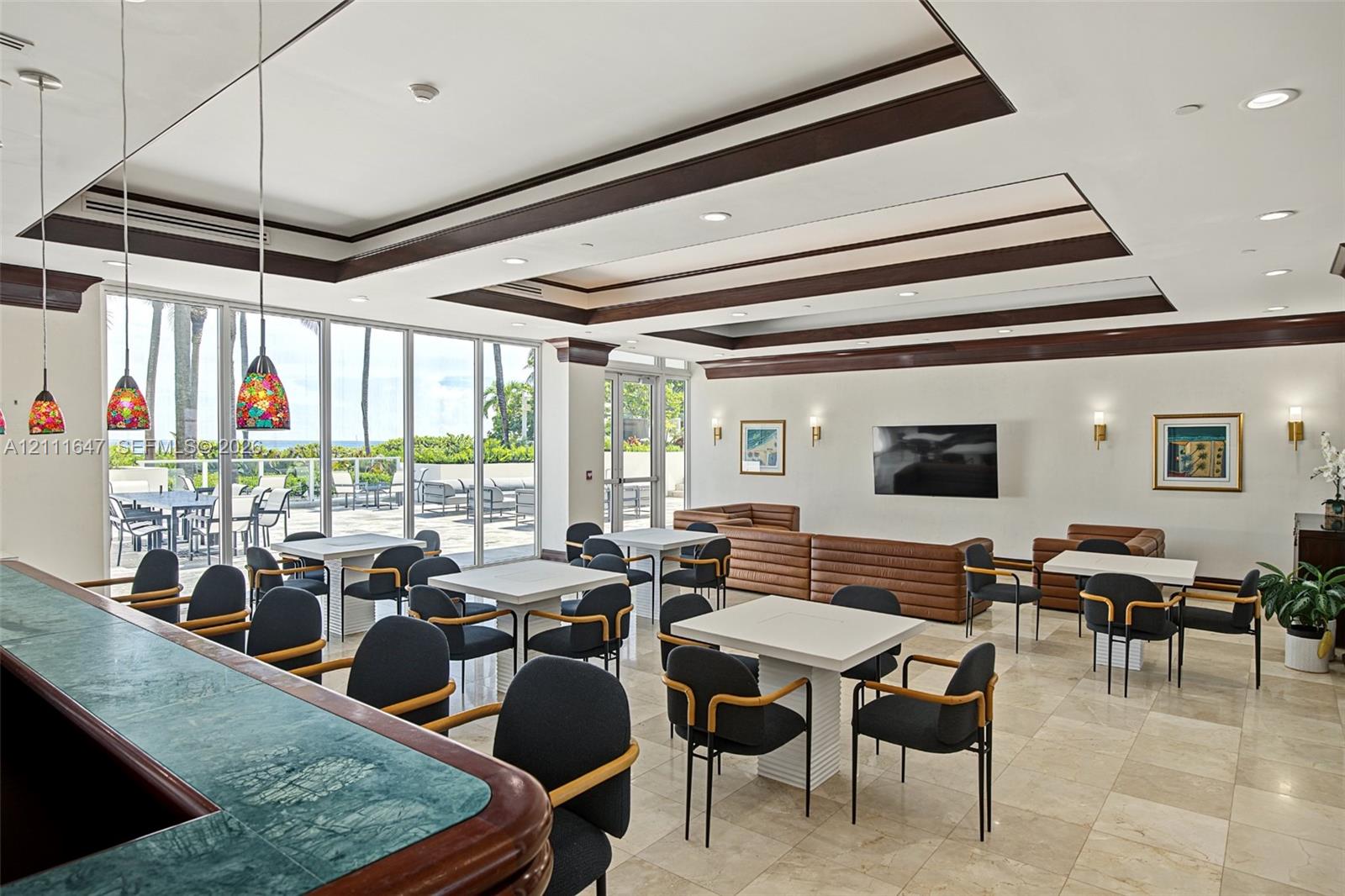 Spacious common area featuring recessed tray ceilings with dark wood trim, tile flooring, built-in stone-top bar, and expansive floor-to-ceiling glass wall panels