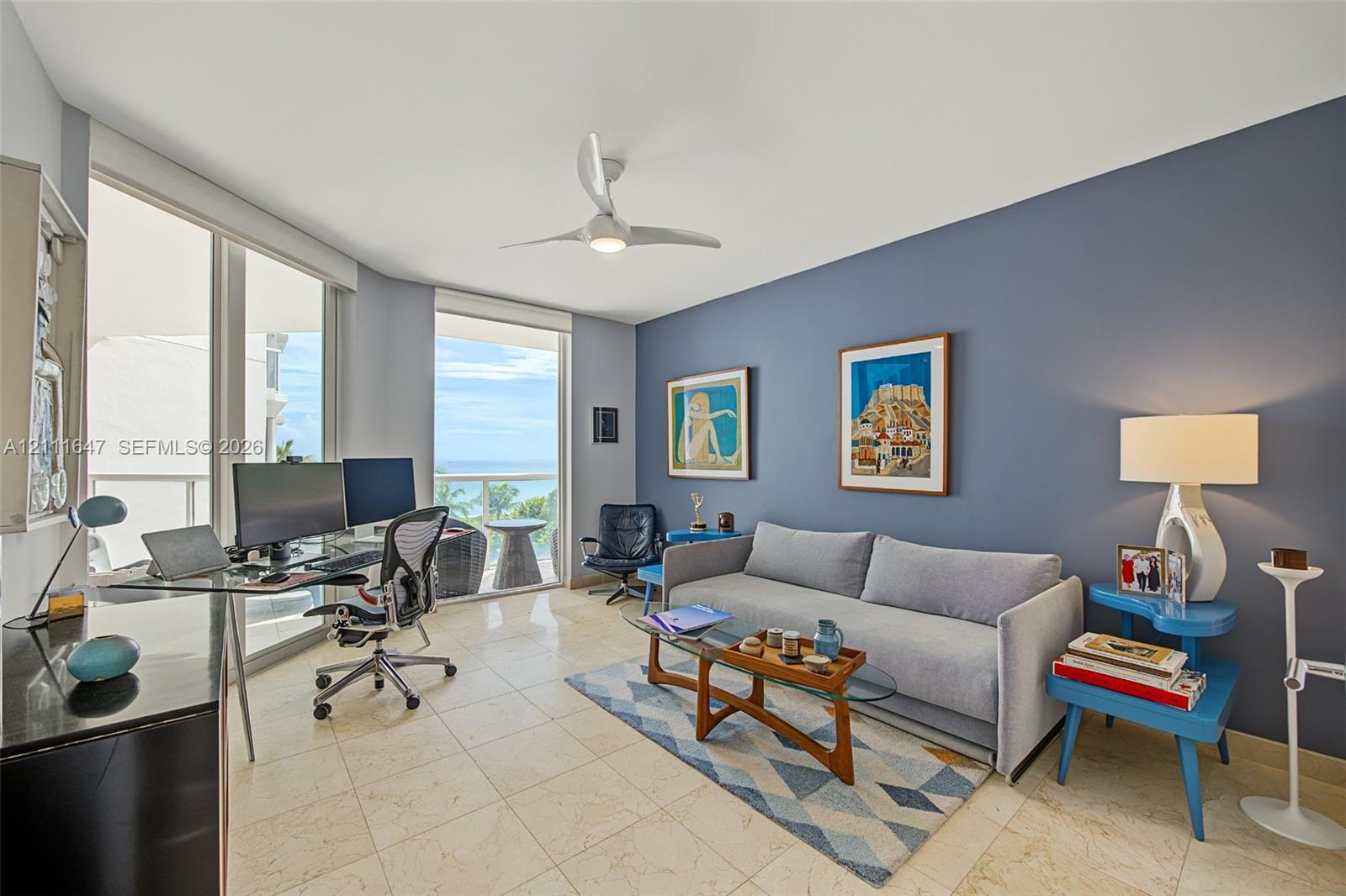 Floor-to-ceiling sliding glass doors, light-toned stone tile flooring, neutral wall palette with a prominent blue accent wall, ceiling fan with integrated lighting, and balcony access