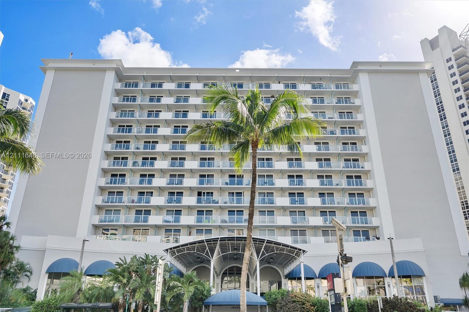 Mid-rise building exterior featuring multiple levels of private balconies with glass railings, a prominent arched entrance canopy, and blue architectural awnings Mid-rise building exterior featuring multiple levels of private balconies with glass railings, a prominent arched entrance canopy, and blue architectural awnings