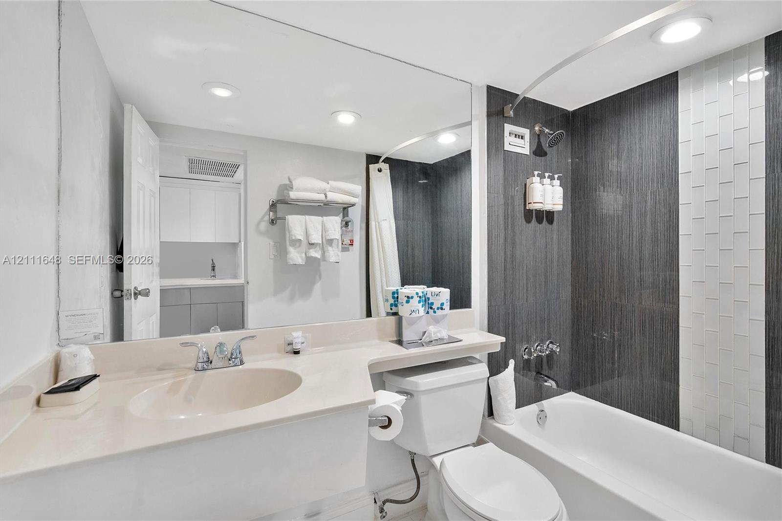 Full-width vanity with an integrated sink, expansive wall-mounted mirror, recessed lighting, and shower area featuring vertical charcoal and white tile surround