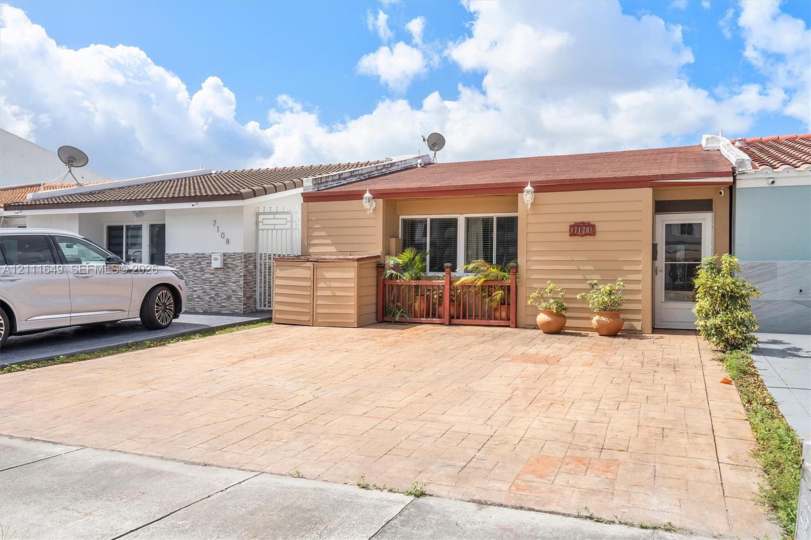 Single-story exterior featuring tan horizontal siding, a paver driveway, a low-profile tiled roof, and an enclosed front porch with a wooden railing
