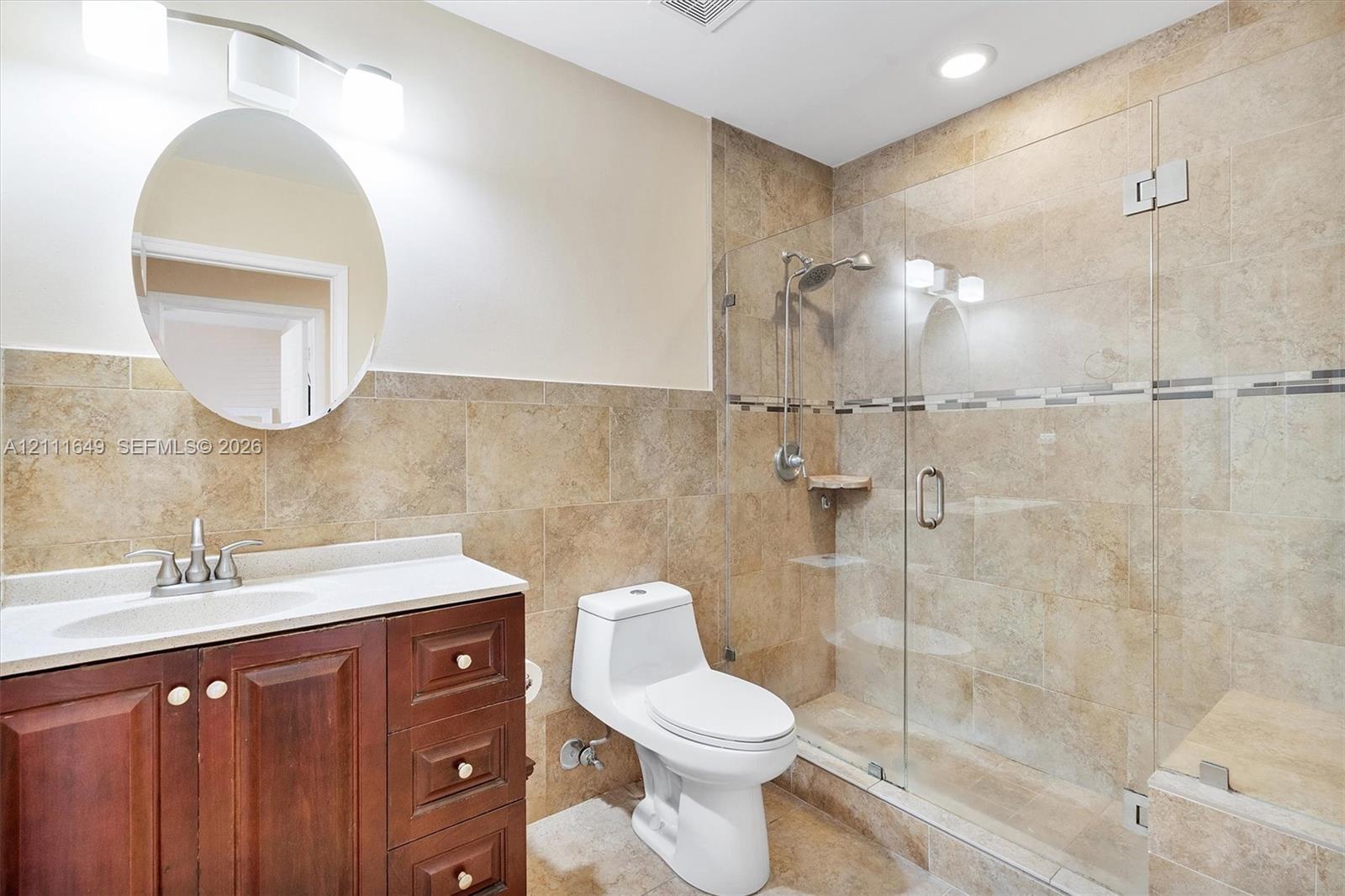 Bathroom featuring a dark wood vanity with a stone countertop, an oval wall-mounted mirror, and a walk-in shower enclosed by glass doors with tiled walls