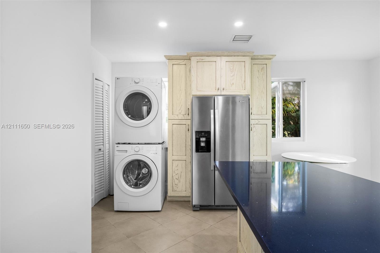 Stacked laundry unit, stainless steel refrigerator, light wood-finish cabinetry, dark solid-surface countertop, and ceramic tile flooring