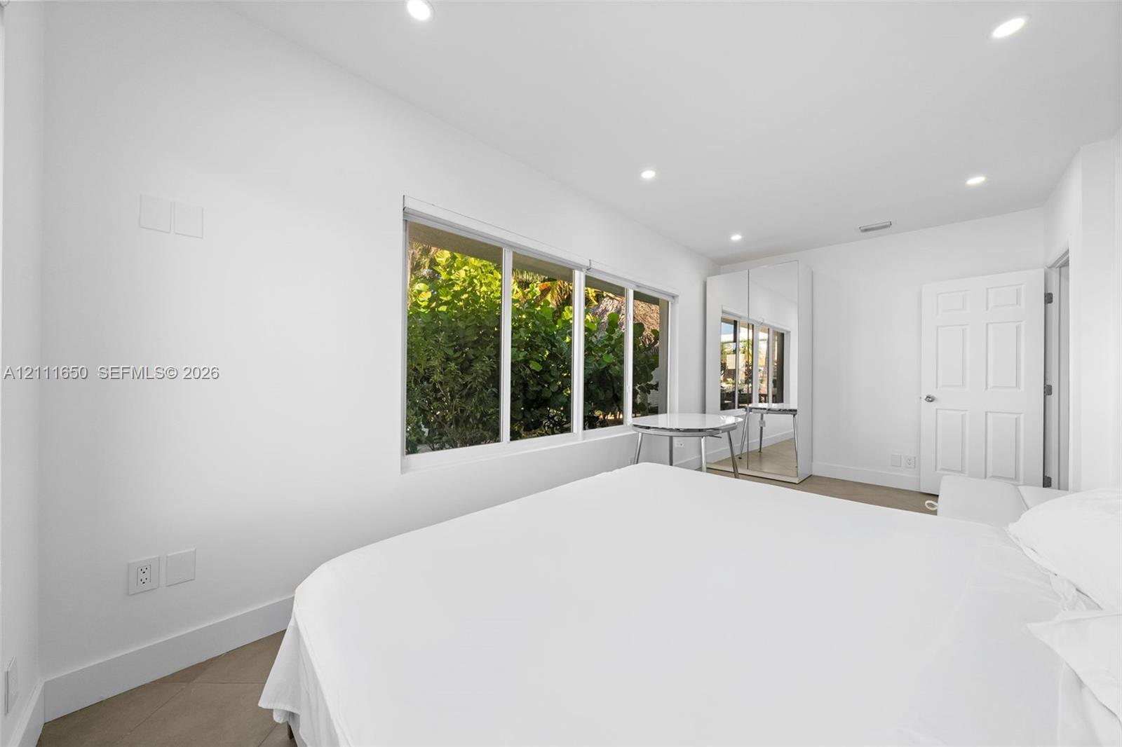 Large horizontal window with exterior foliage, neutral-toned flooring, recessed ceiling lighting, and full-height mirrored built-in closet doors
