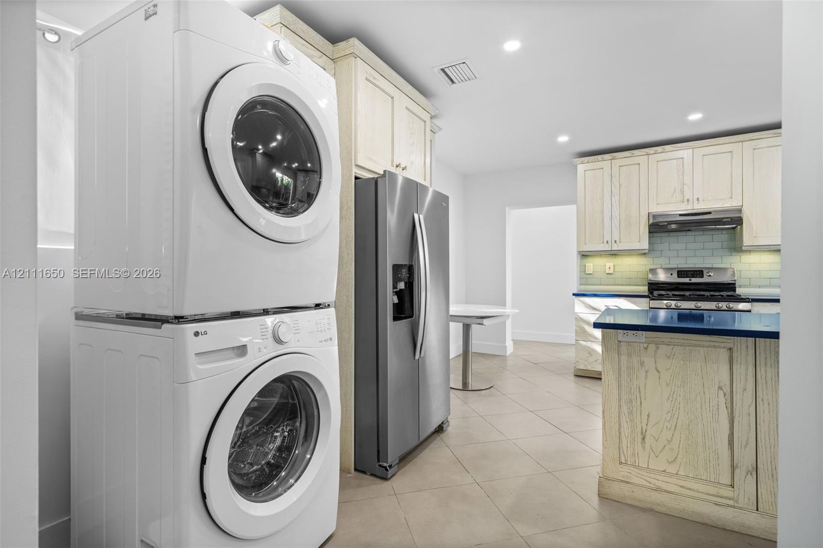 Stacked front-loading washer and dryer, stainless steel French-door refrigerator, tile flooring, custom cabinetry, and stone-surface countertops