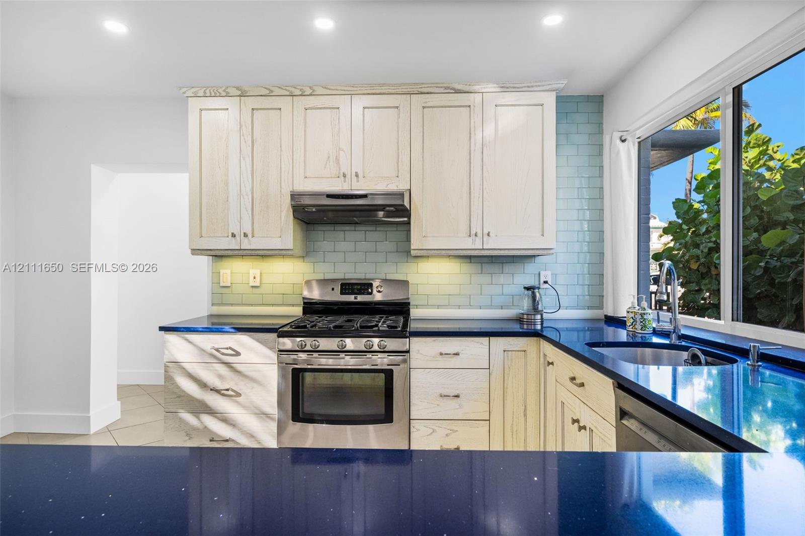 Kitchen featuring light-toned wood-finish cabinetry, contrasting dark countertops, a stainless steel gas range with overhead vent, and blue subway tile backsplash