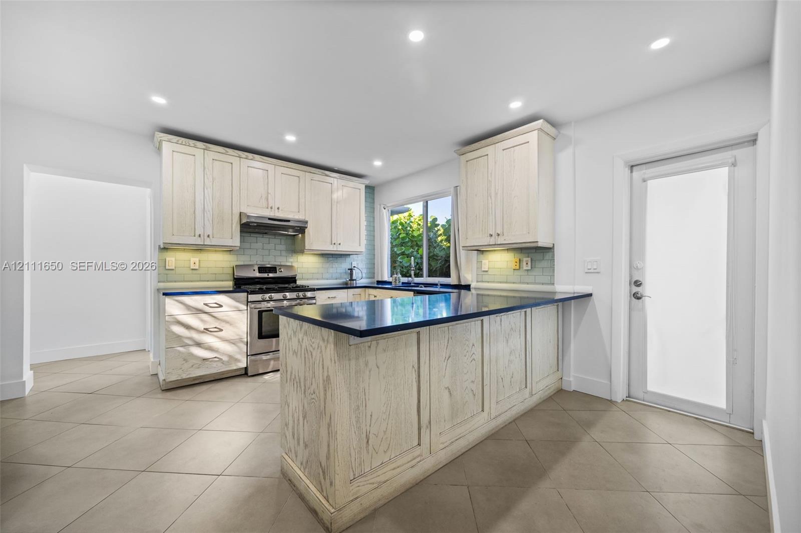 Kitchen featuring light-toned wood cabinetry, dark solid-surface countertops, a subway tile backsplash, recessed lighting, and tile flooring