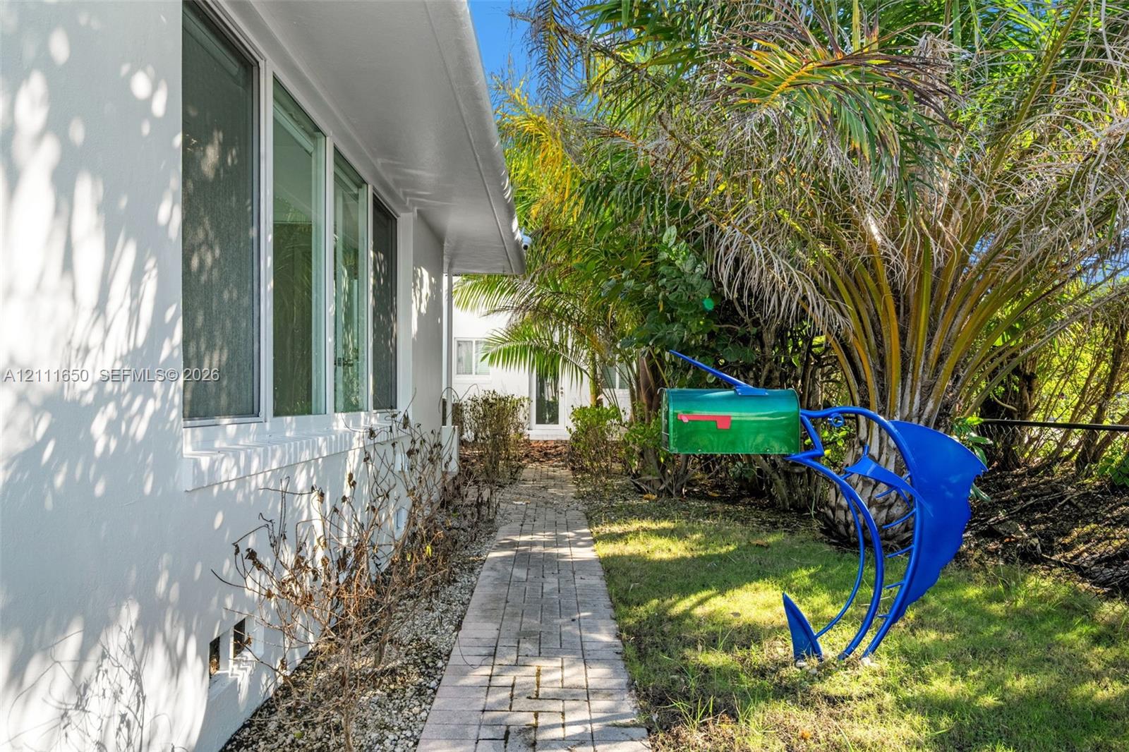 Exterior side profile featuring smooth-finish white stucco walls, multi-pane windows, and a paved brick walkway bordered by mature palms and foliage