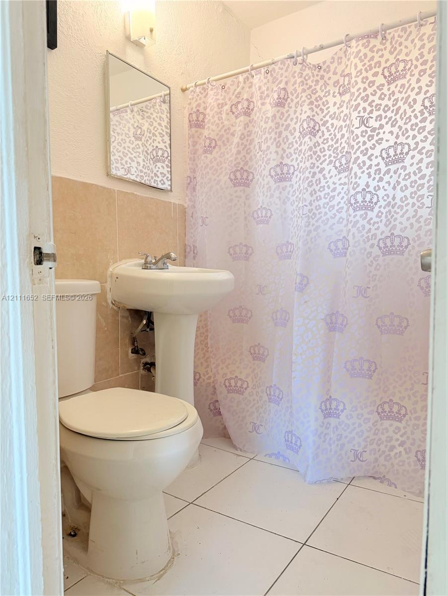 Bathroom featuring white floor tiling, pedestal sink with dual-handle faucet, wall-mounted rectangular mirror, and neutral-toned wall tile surround