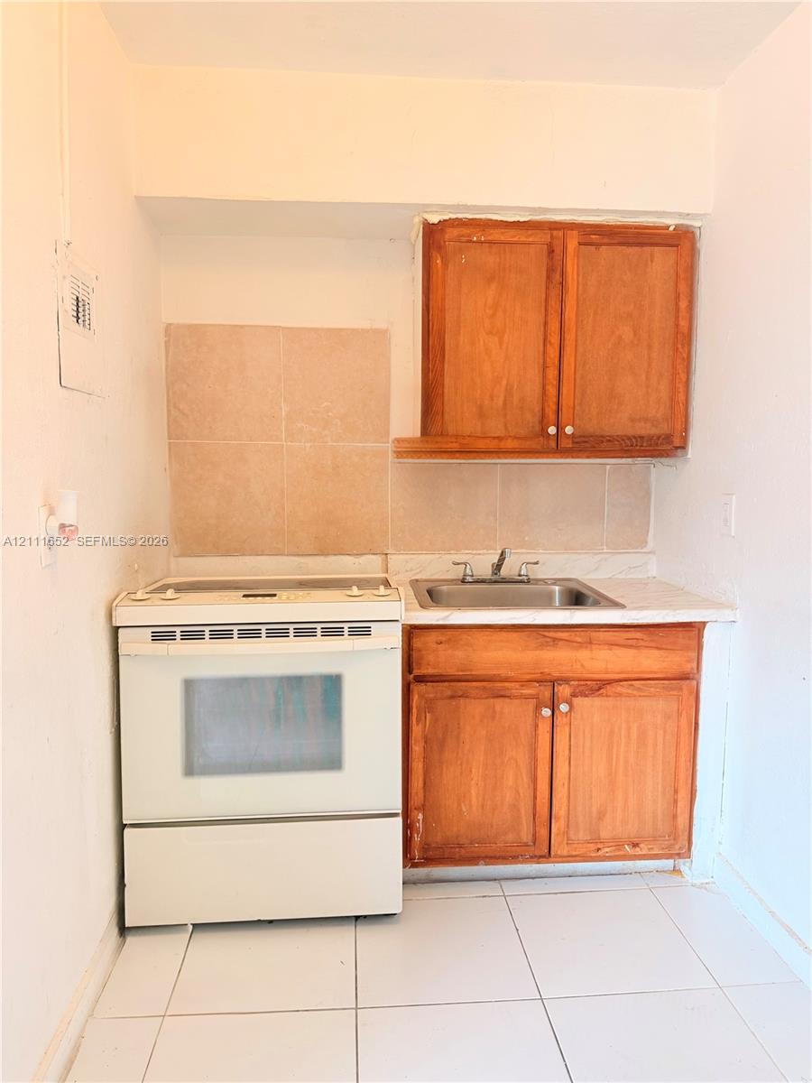 Kitchenette featuring white porcelain floor tiles, wood-finish cabinetry, a single-basin stainless steel sink, a white electric range, and a tiled backsplash