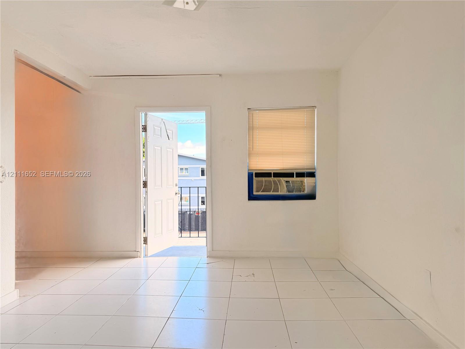 Interior space featuring white ceramic tile flooring, a window-integrated air conditioning unit, and a doorway opening to a metal-railed exterior balcony