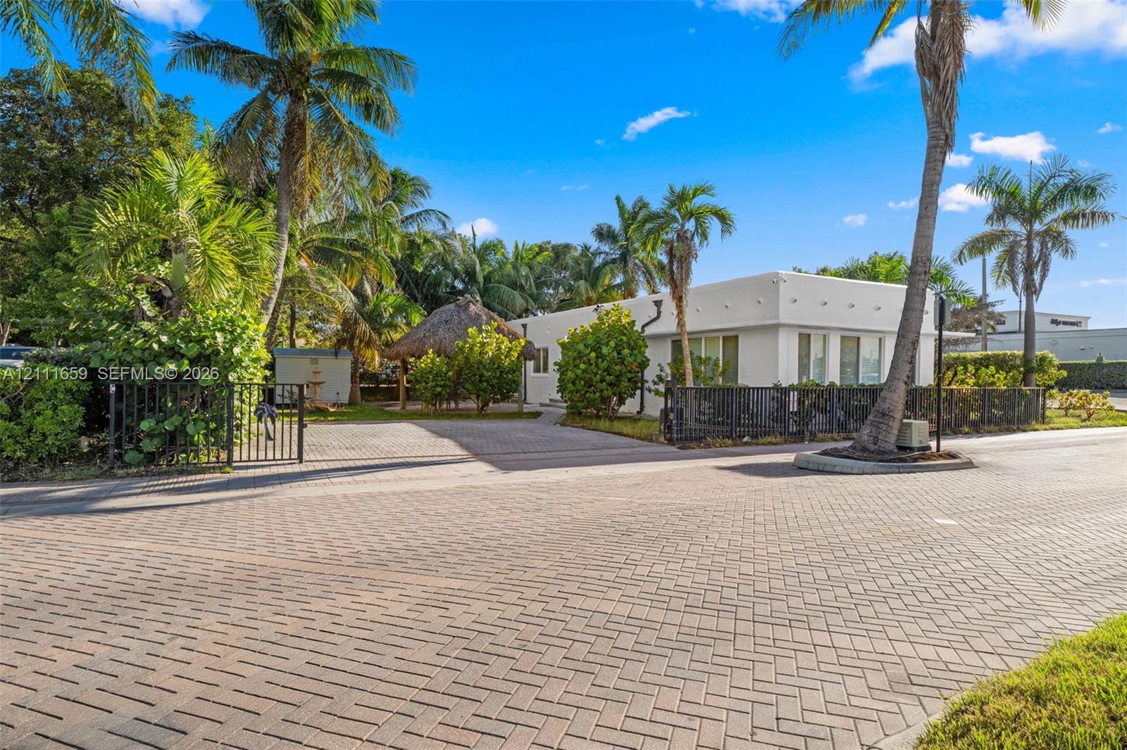 Exterior featuring a white flat-roof facade, a herringbone-patterned brick paver driveway, an outdoor thatched-roof structure, and perimeter metal fencing