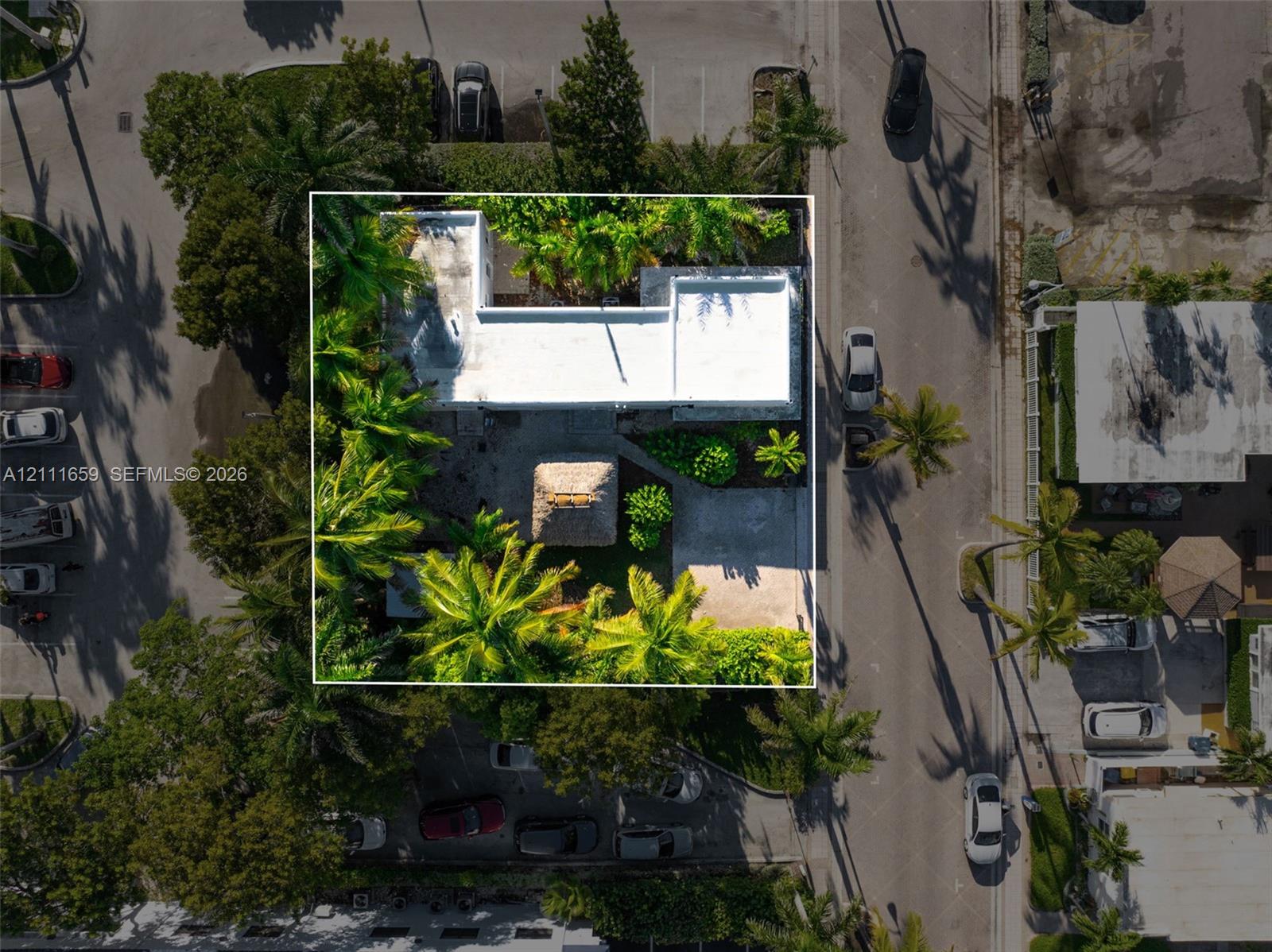 Aerial perspective of a property featuring a flat-roof residence, a detached thatch-roof structure, concrete hardscaping, and mature palm trees