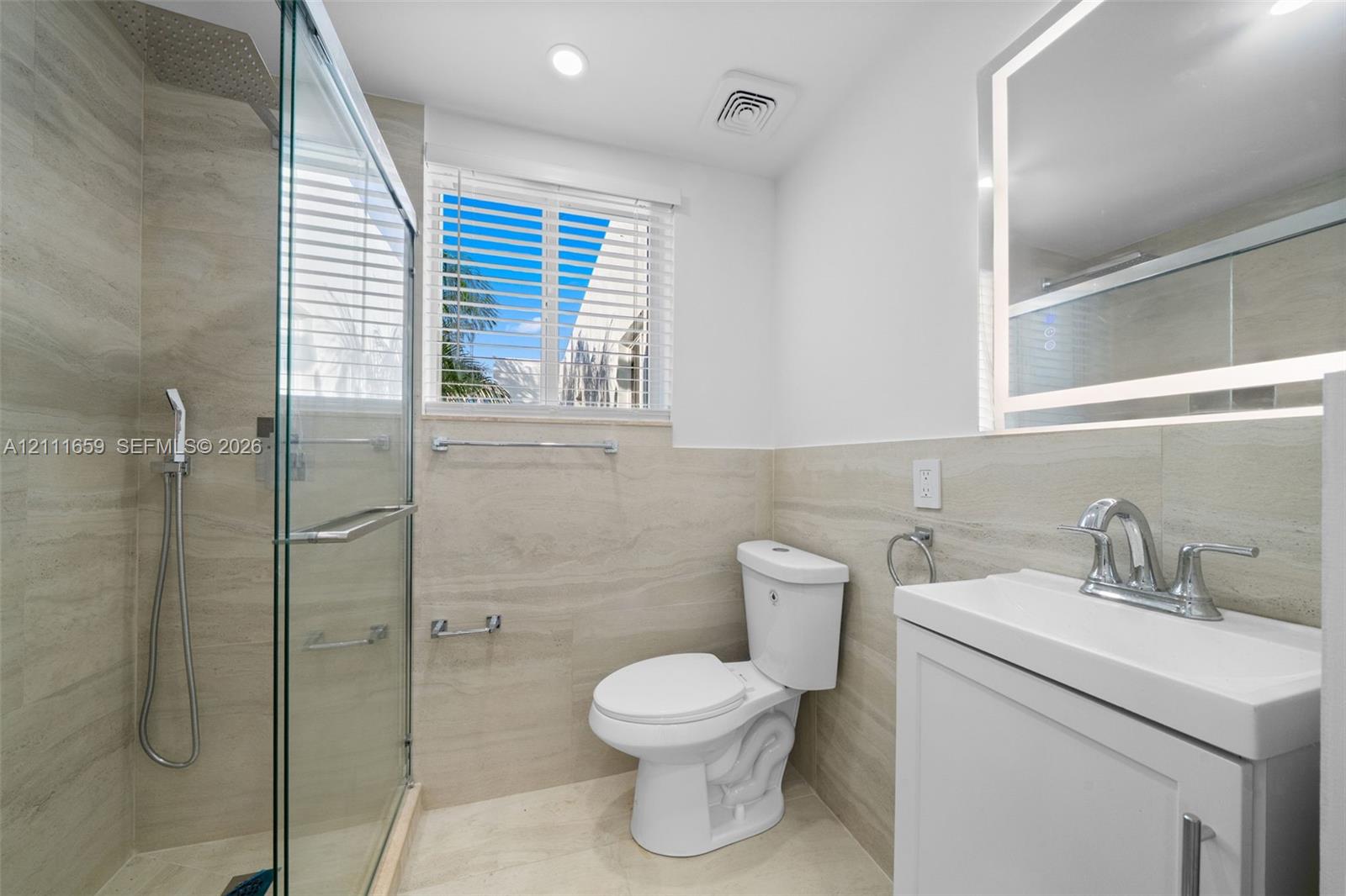 Bathroom featuring walk-in shower with glass enclosure, stone-finish wall tile, illuminated vanity mirror, and white cabinetry