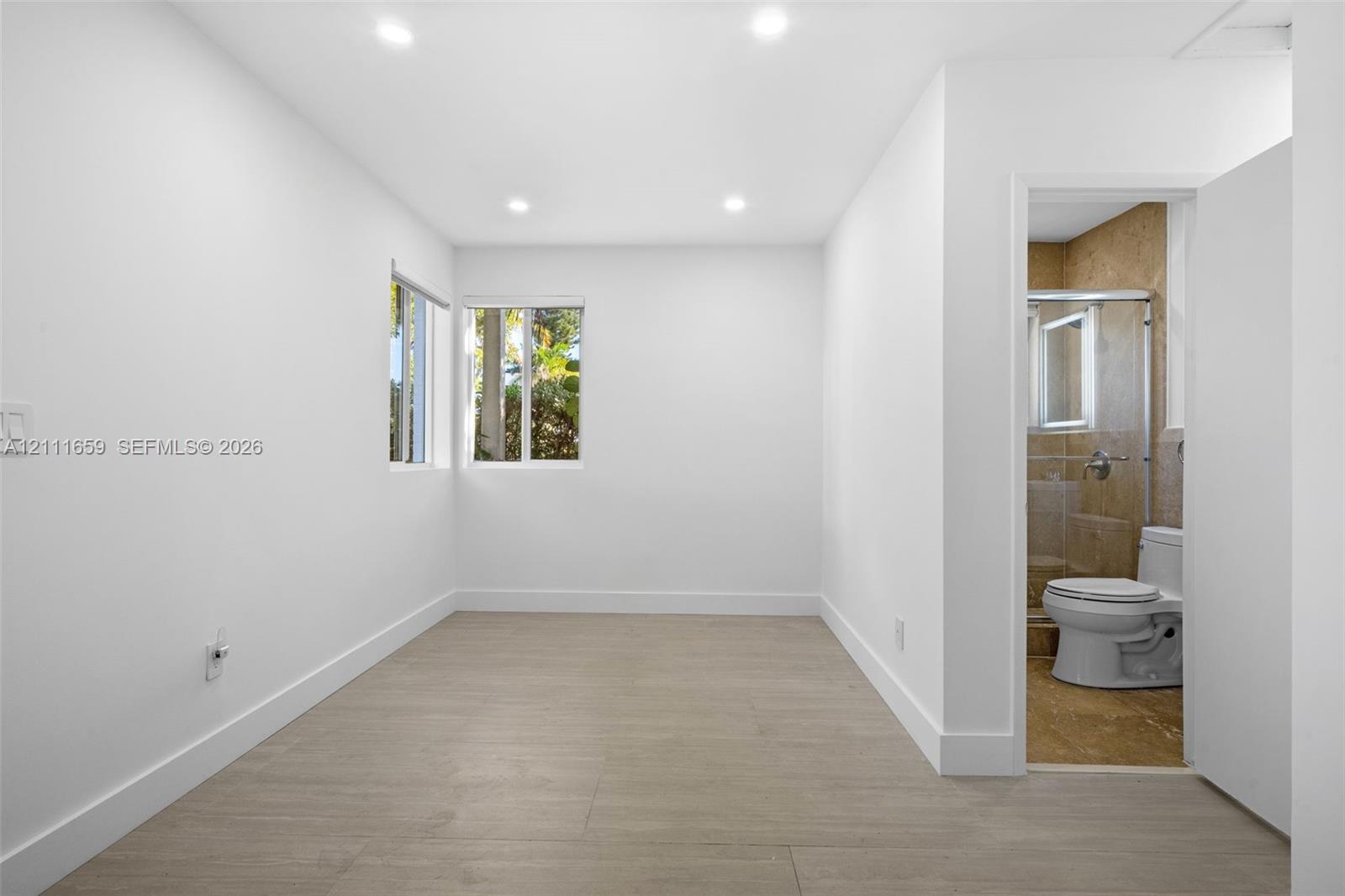 Interior space featuring wood-finish flooring, recessed lighting, and dual windows, with an attached bathroom showcasing tile wall surfacing, a glass-enclosed shower, and a vitreous china toilet