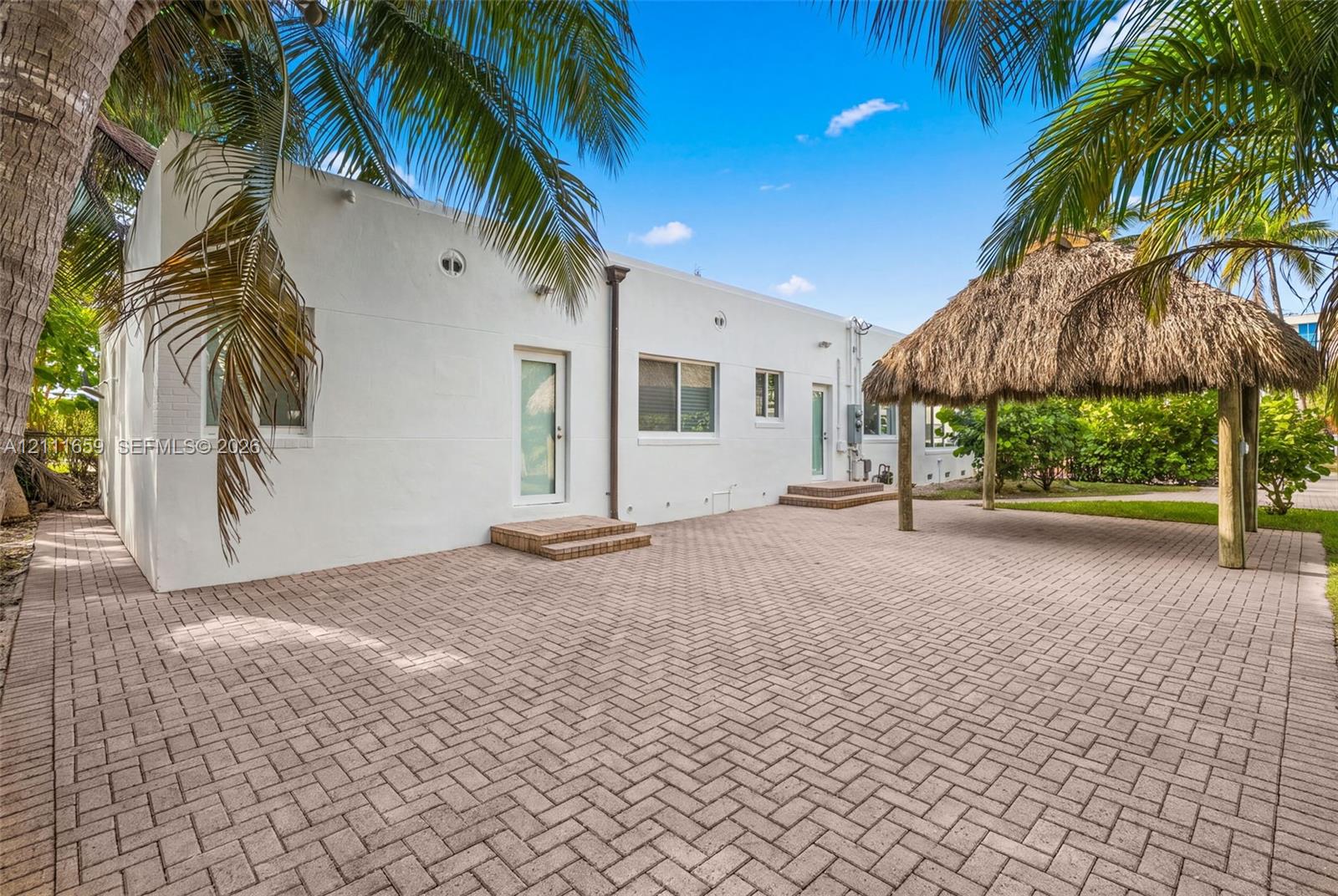 Rear exterior featuring a herringbone-pattern paver patio, thatched-roof tiki hut, white stucco facade, and mature palm trees