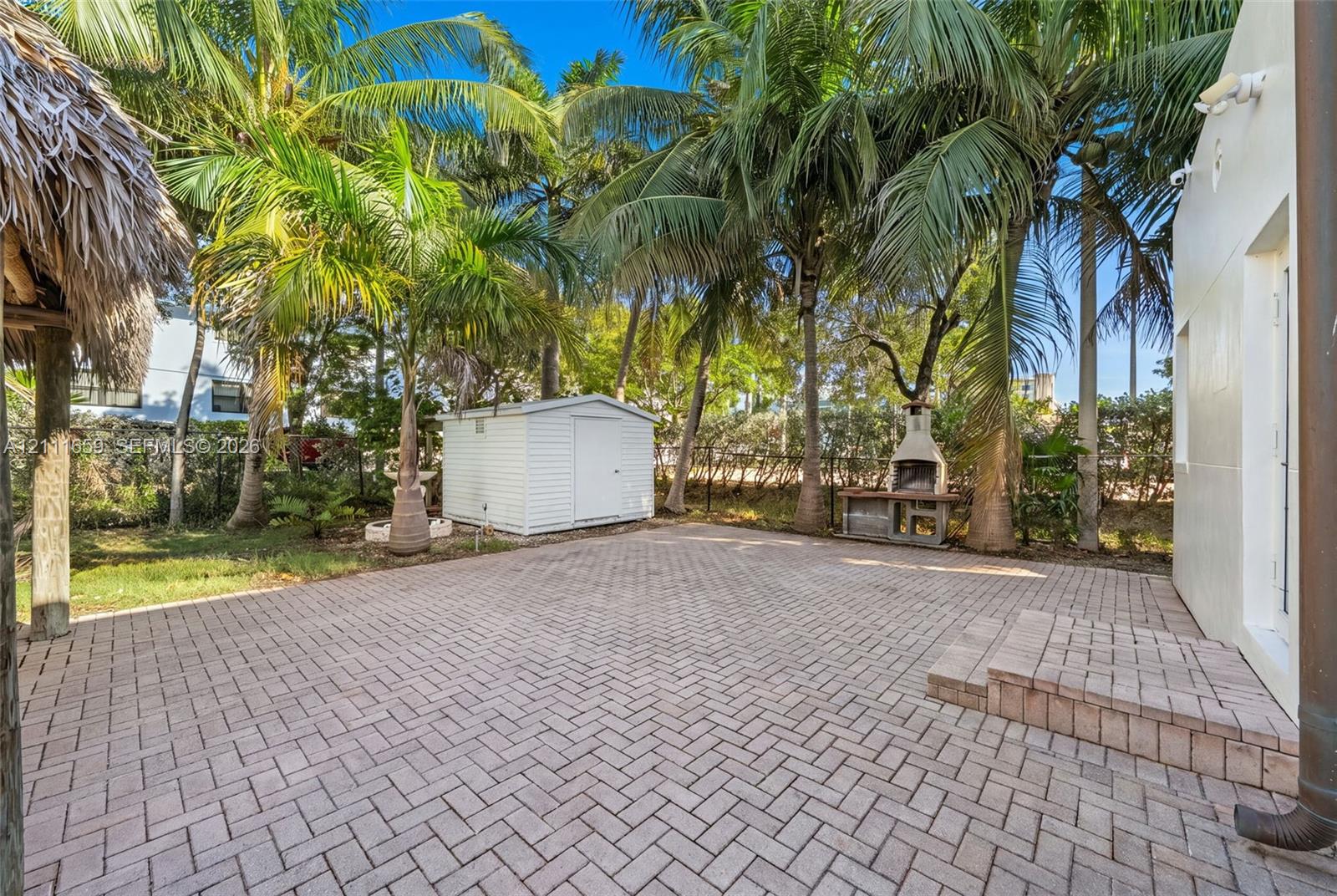 Outdoor space featuring a herringbone-patterned paver patio, permanent masonry outdoor fireplace, detached storage structure, and mature palm trees