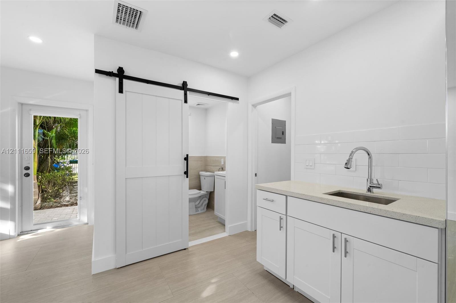 White shaker-style cabinetry with neutral solid-surface countertop, subway tile backsplash, stainless steel under-mount sink, and sliding barn-style door