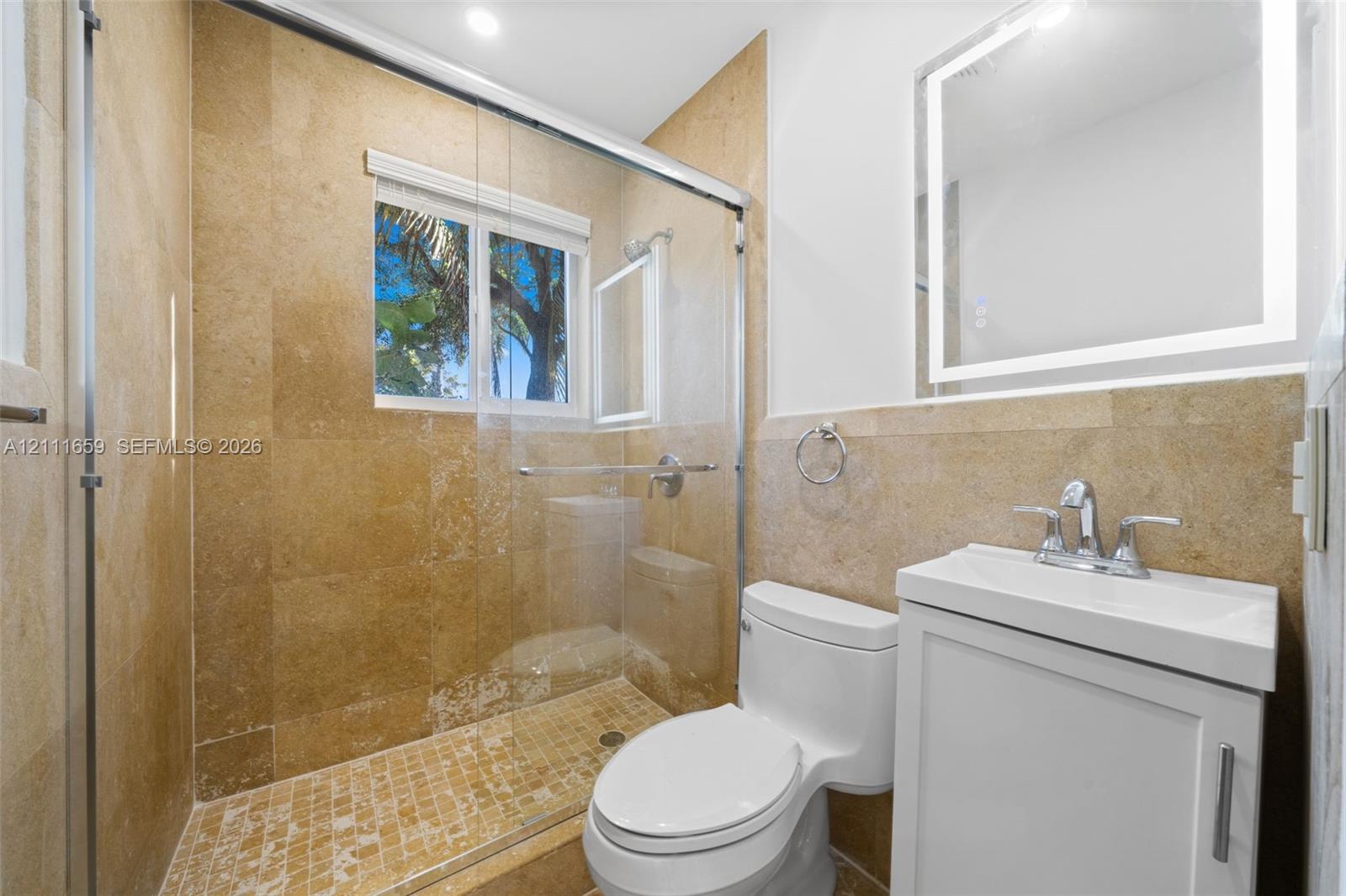 Bathroom featuring stone tile shower walls with an integrated glass enclosure, custom-lit vanity mirror, white cabinetry, and mosaic tile shower flooring
