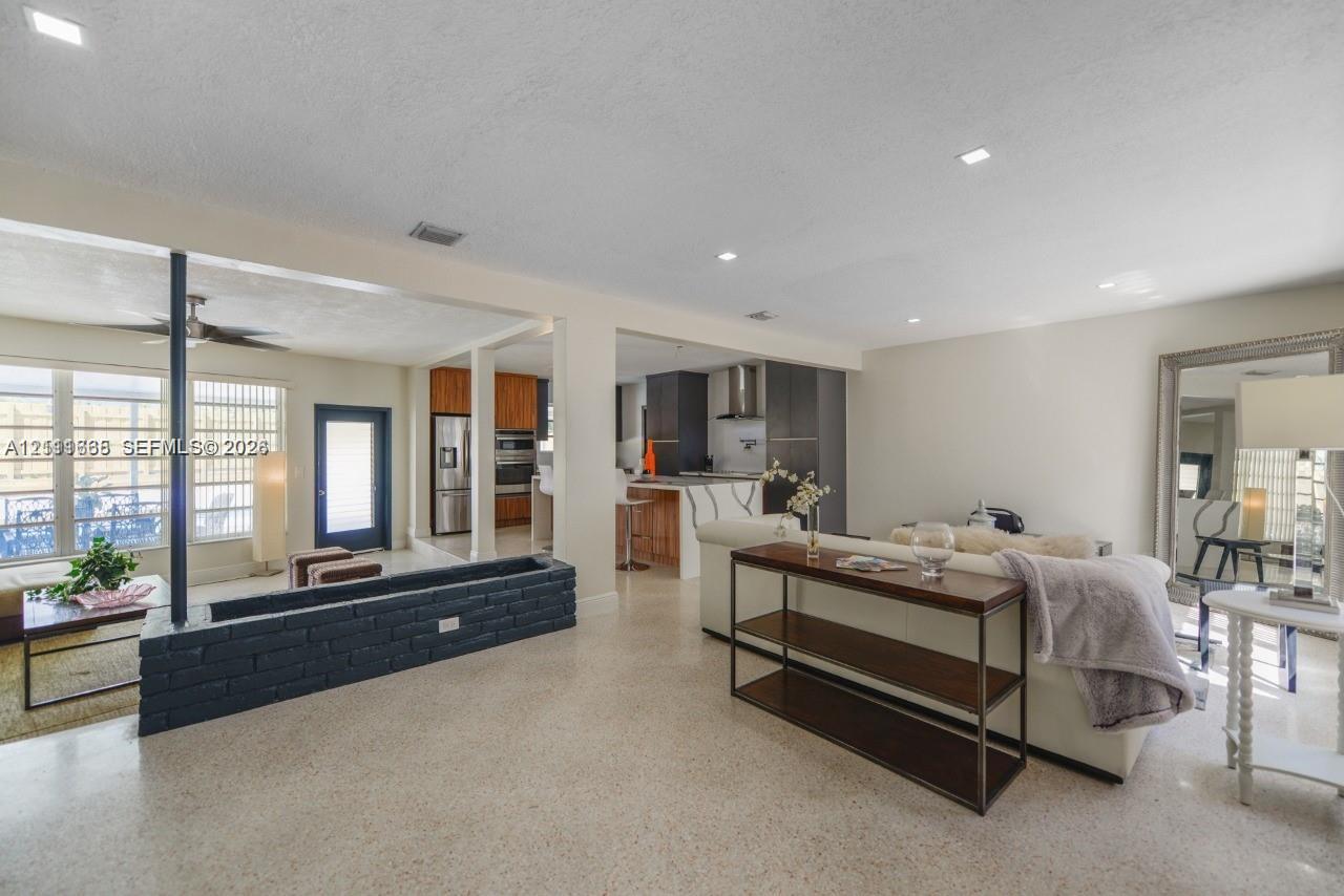 Open-concept interior featuring light-toned stone flooring, a painted brick structural partition, recessed lighting, and a kitchen equipped with dark cabinetry and a central island