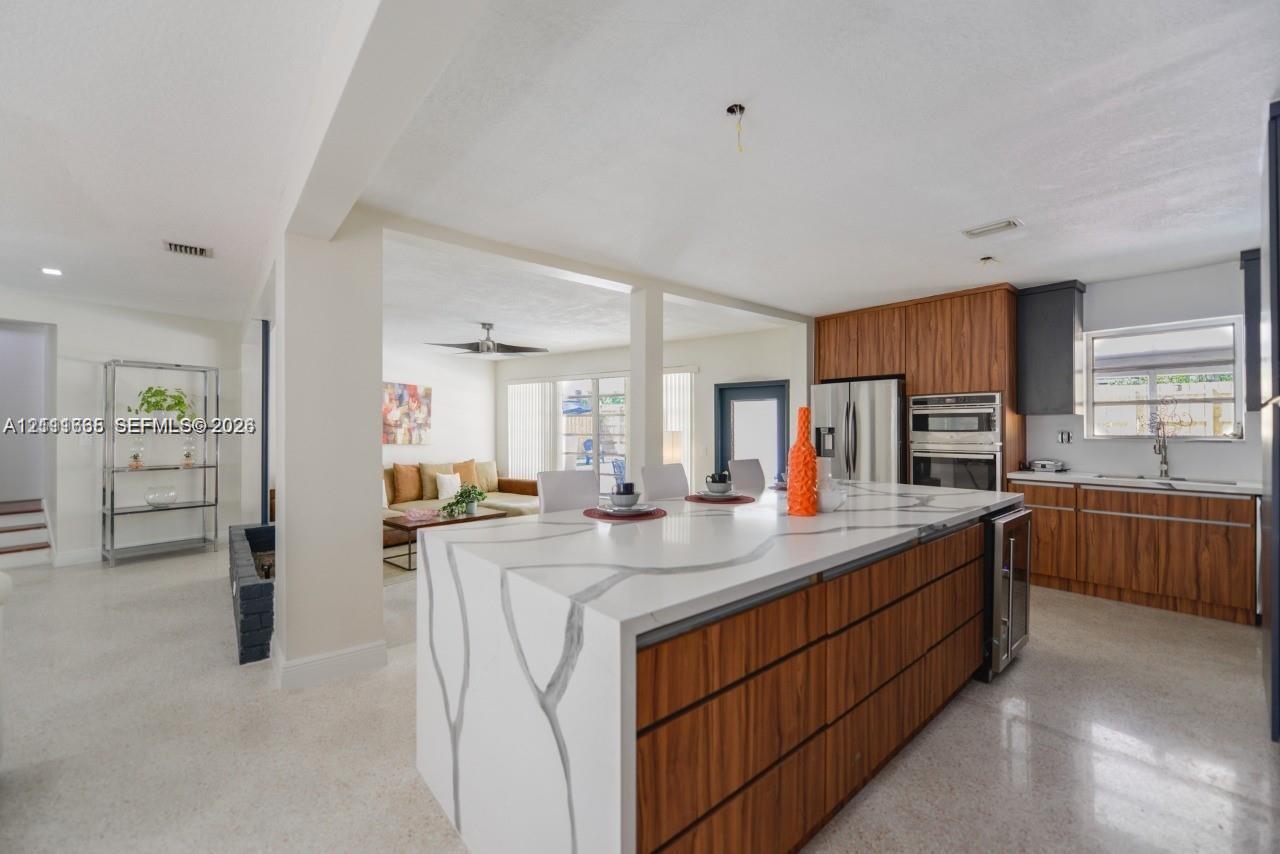 Kitchen featuring a central island with a stone-finish countertop and waterfall edge, wood-finish cabinetry, stainless steel built-in wall oven, and light-colored terrazzo flooring