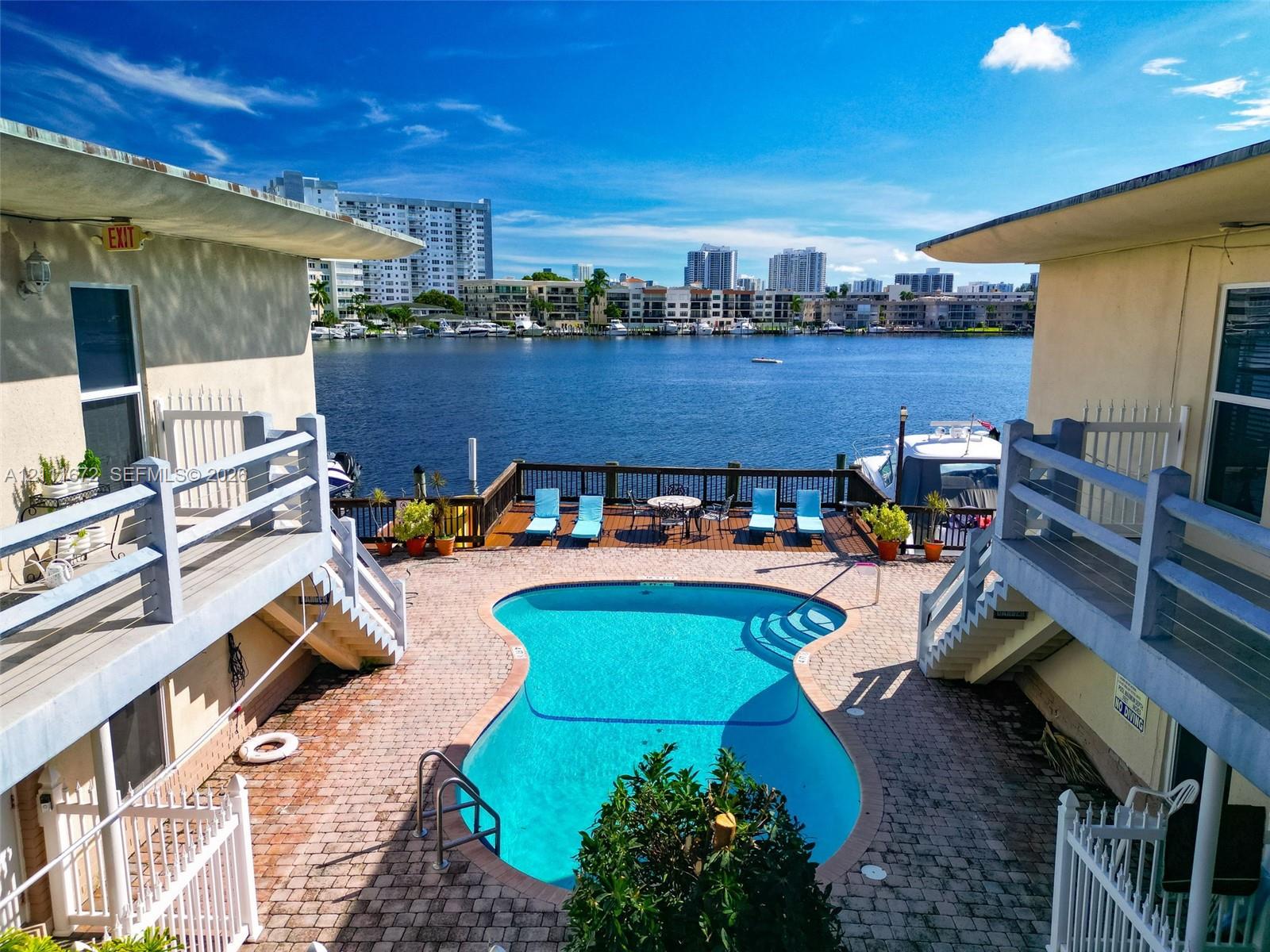 Waterfront swimming pool surrounded by a brick paver deck, wooden dock, and balcony railings