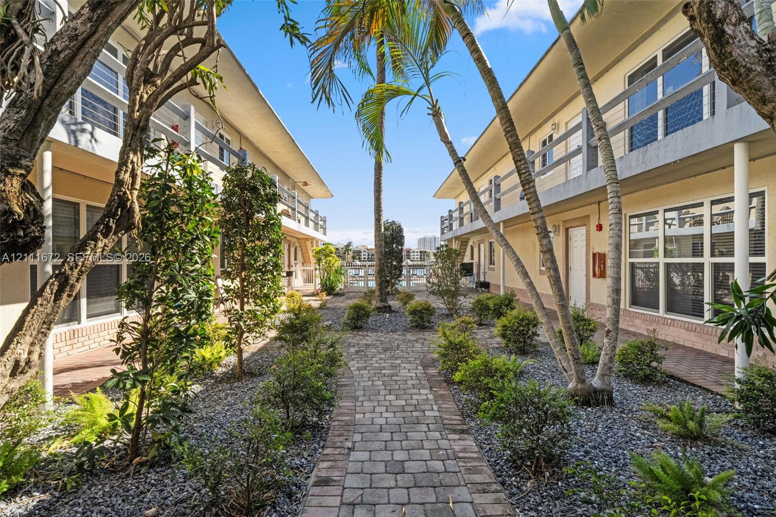 Paver stone walkway lined with mature palm trees and manicured shrubbery, featuring multi-story structures with tiered balconies and exterior staircases Paver stone walkway lined with mature palm trees and manicured shrubbery, featuring multi-story structures with tiered balconies and exterior staircases