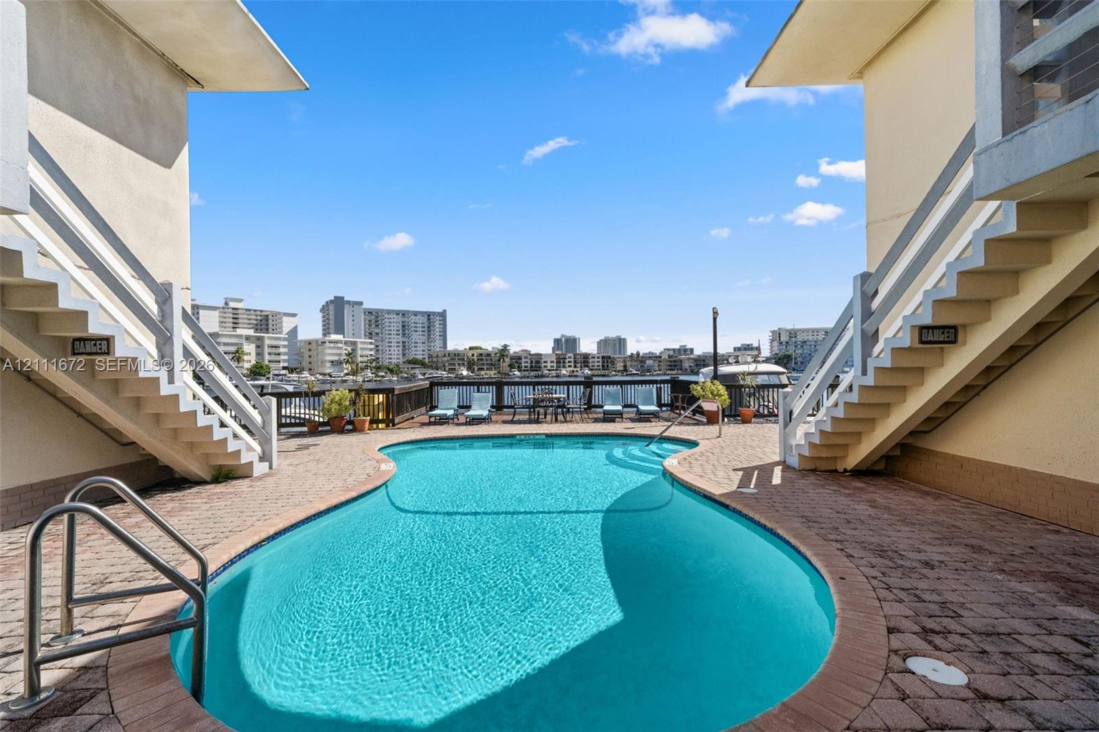 Curved swimming pool featuring a brick-paver deck, stainless steel entry rails, and a wooden perimeter railing overlooking the water Curved swimming pool featuring a brick-paver deck, stainless steel entry rails, and a wooden perimeter railing overlooking the water