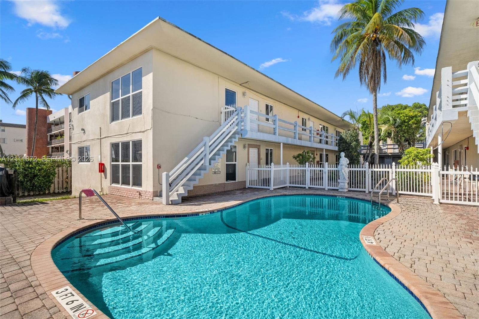 In-ground swimming pool with stone-paver decking, white picket fencing, and two-story building exterior featuring exterior staircases In-ground swimming pool with stone-paver decking, white picket fencing, and two-story building exterior featuring exterior staircases