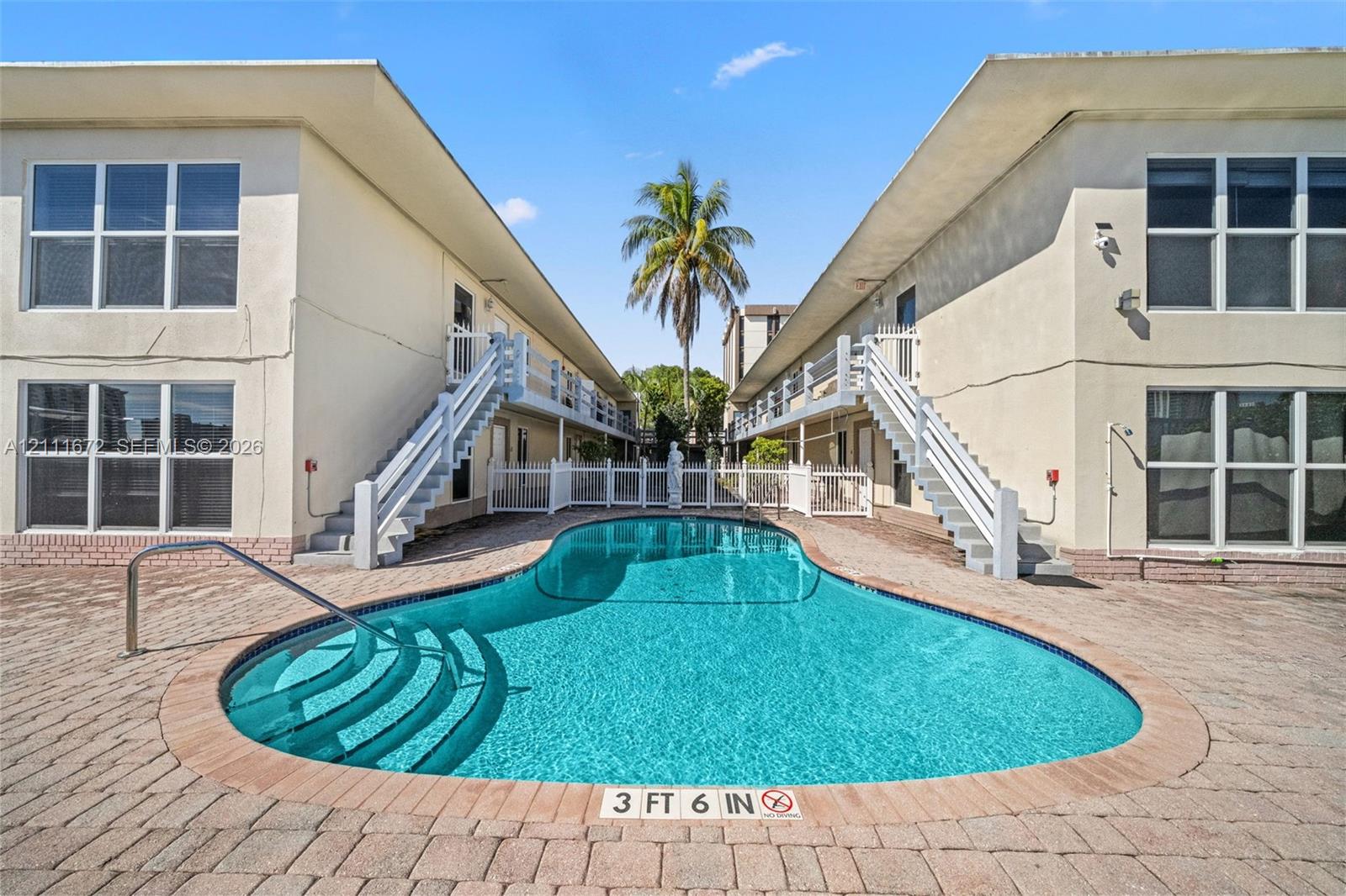 In-ground swimming pool featuring curved entry steps, surrounded by a brick paver deck, dual staircases with white railings, and multi-pane window configurations In-ground swimming pool featuring curved entry steps, surrounded by a brick paver deck, dual staircases with white railings, and multi-pane window configurations