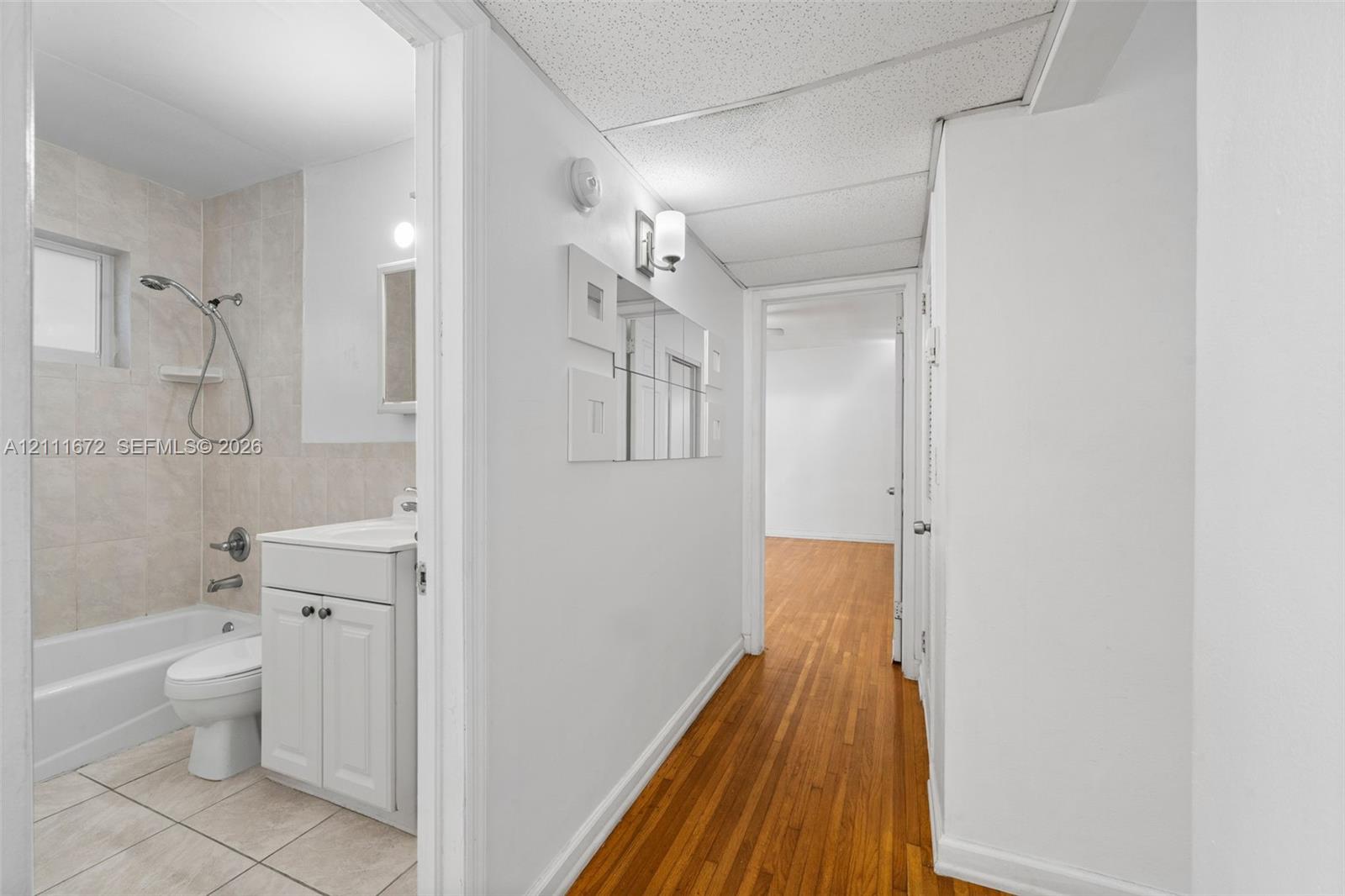 Bathroom featuring a white vanity cabinet, tiled tub surround, and tile flooring, paired with wood-finish flooring in the hallway
