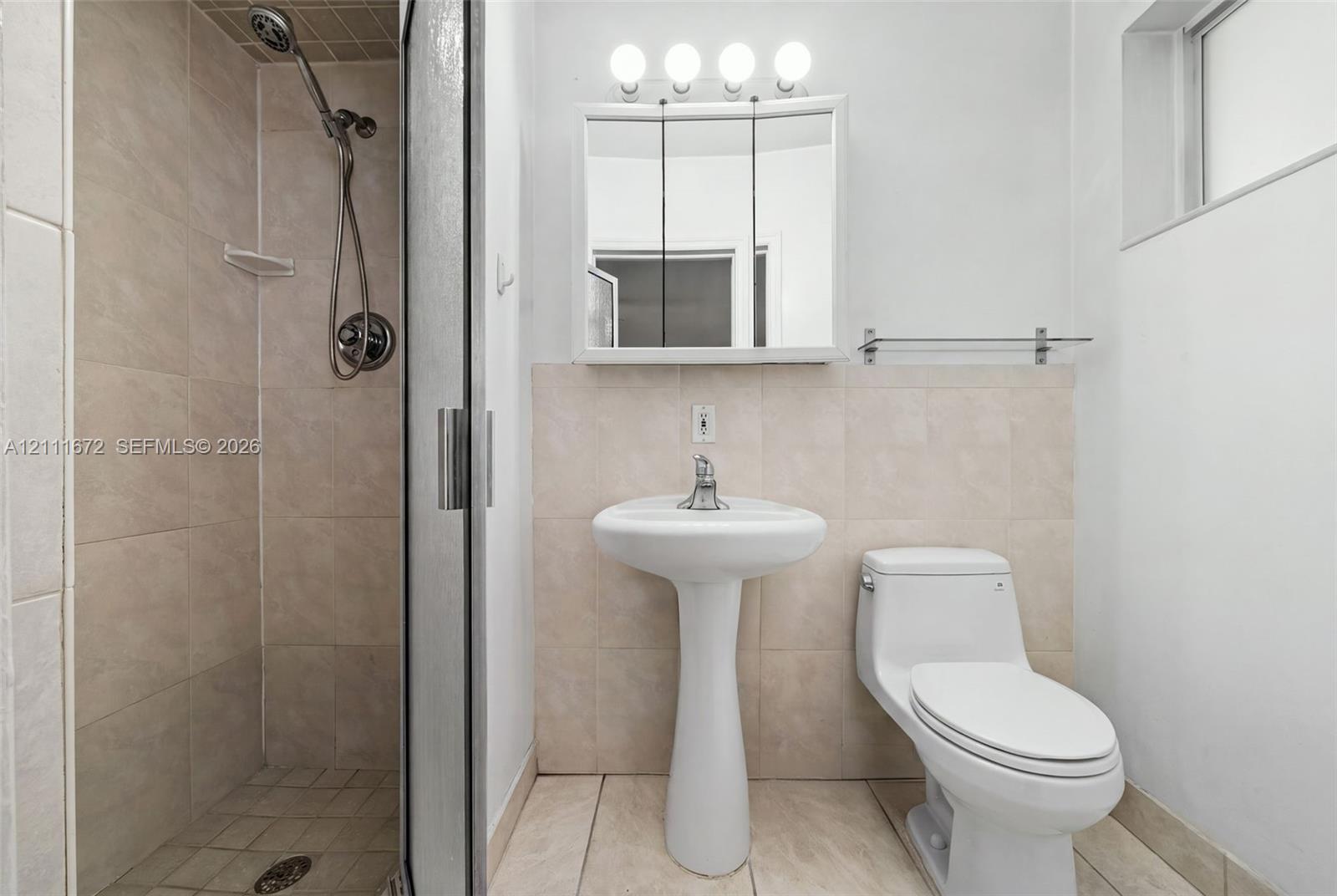 Bathroom featuring a tiled stall shower with a handheld fixture, pedestal sink, mirrored medicine cabinet with integrated lighting, and tile flooring