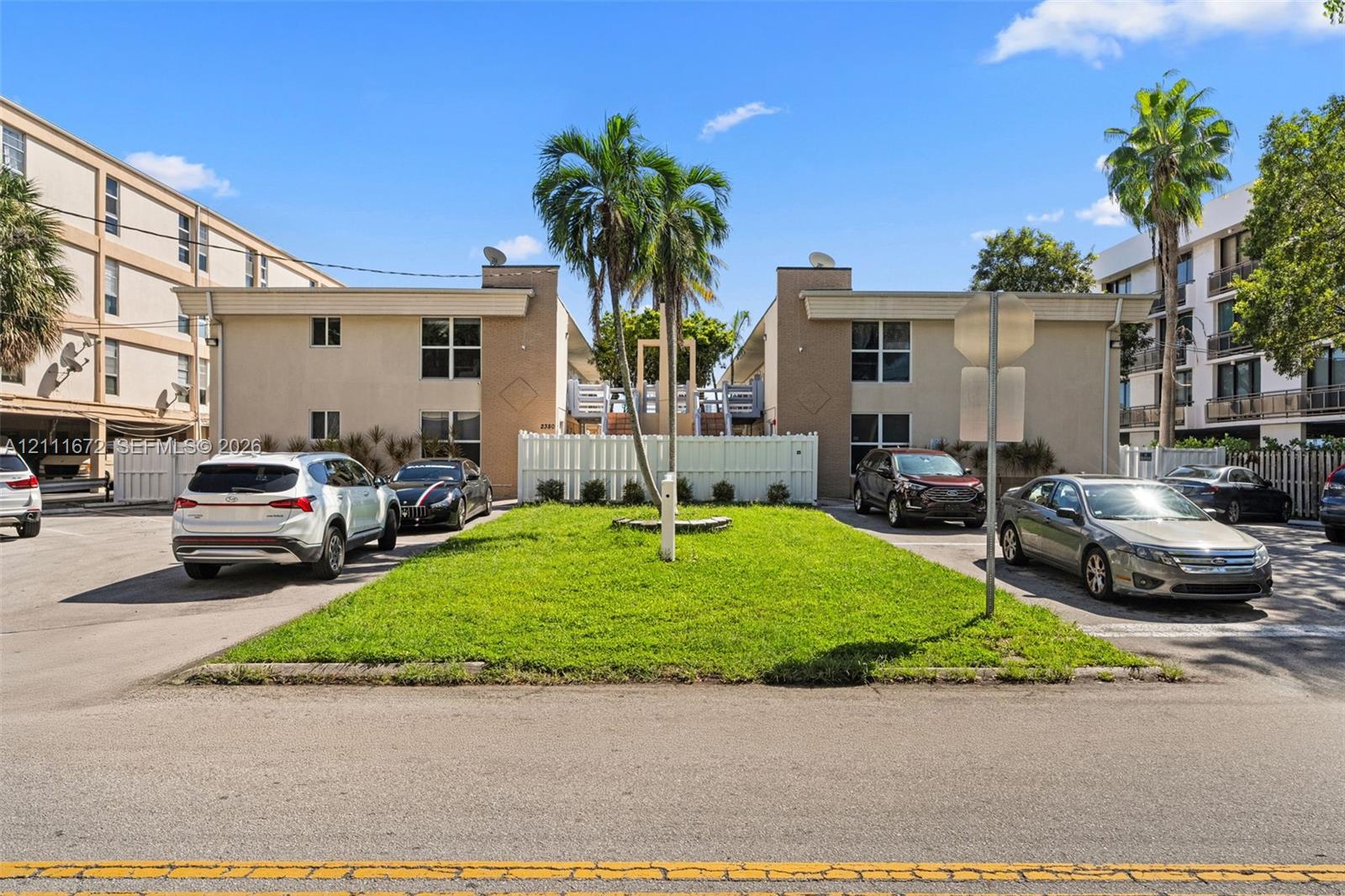 Two-story multi-family exterior featuring stucco facade, surface-level parking area, and manicured center lawn with palm trees Two-story multi-family exterior featuring stucco facade, surface-level parking area, and manicured center lawn with palm trees