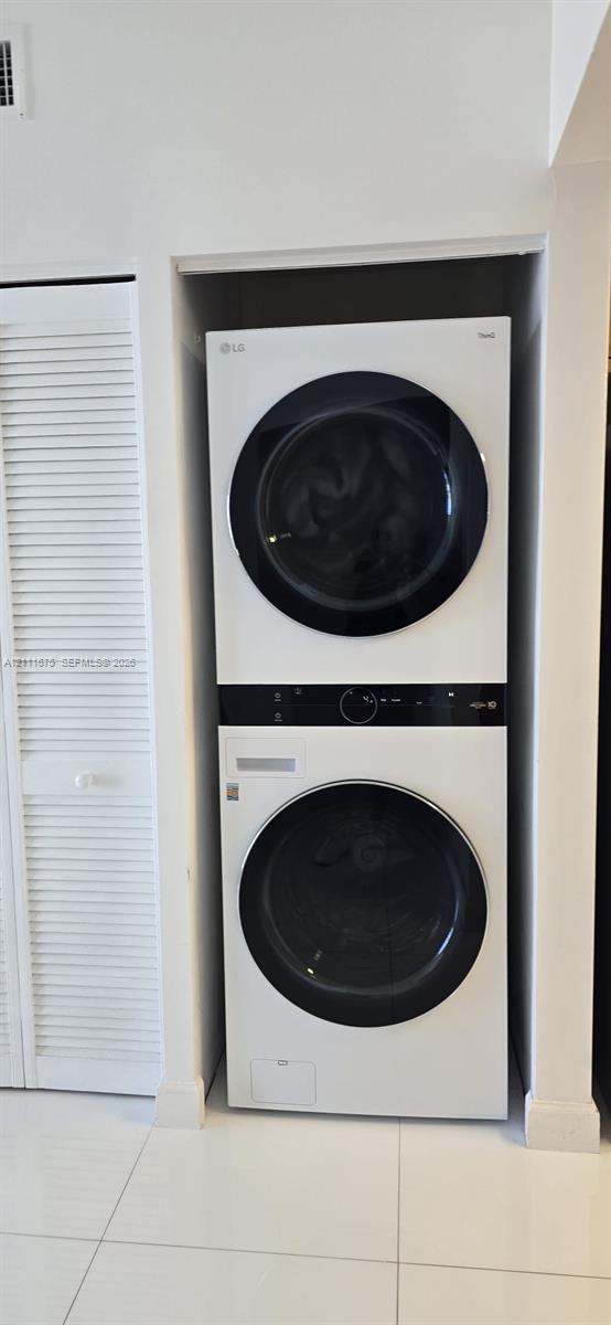 Stacked laundry unit featuring a white finish with front-loading glass-panel doors, complemented by white louvered closet doors and large-format tile flooring