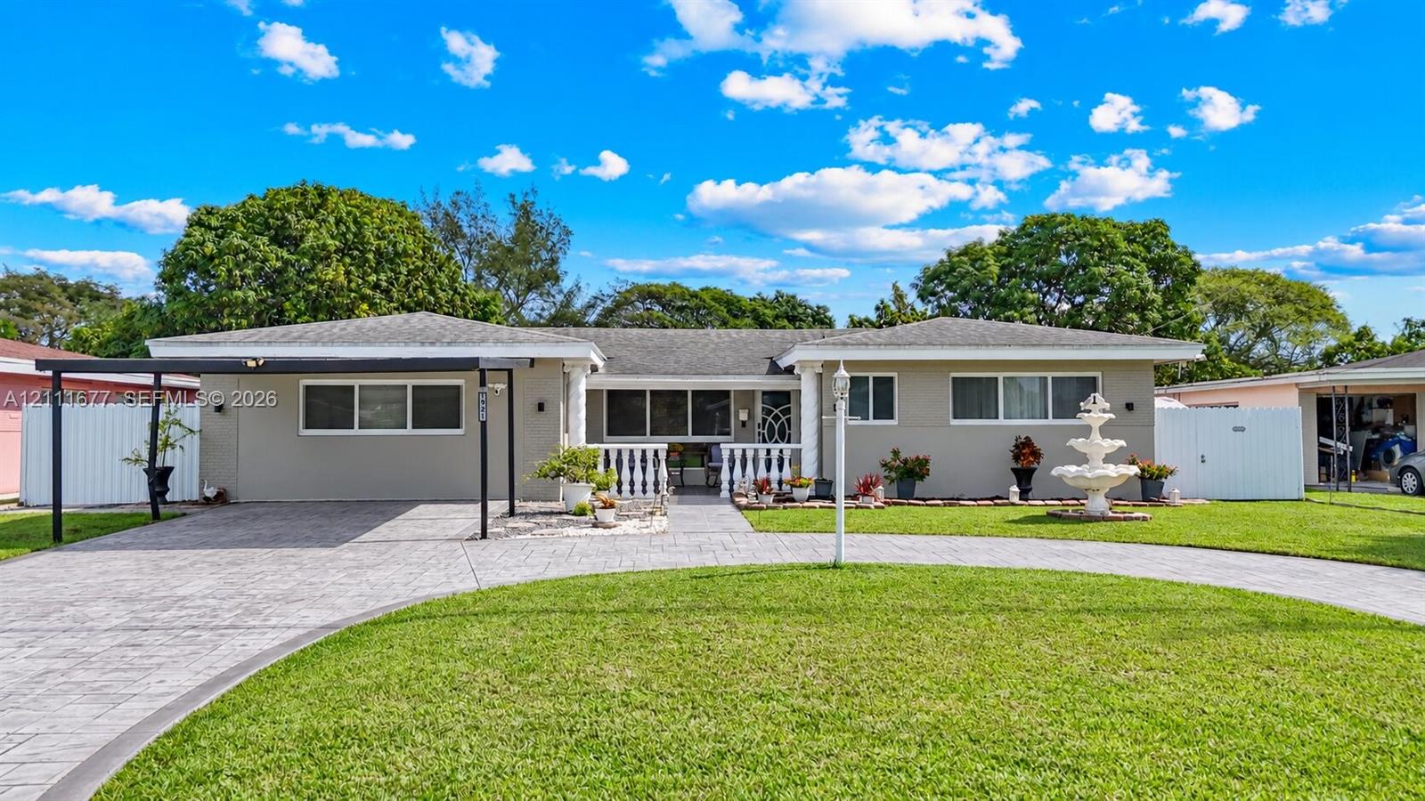 Single-story exterior featuring a circular paver driveway, covered carport, decorative fountain, and white balustrade porch railing Single-story exterior featuring a circular paver driveway, covered carport, decorative fountain, and white balustrade porch railing